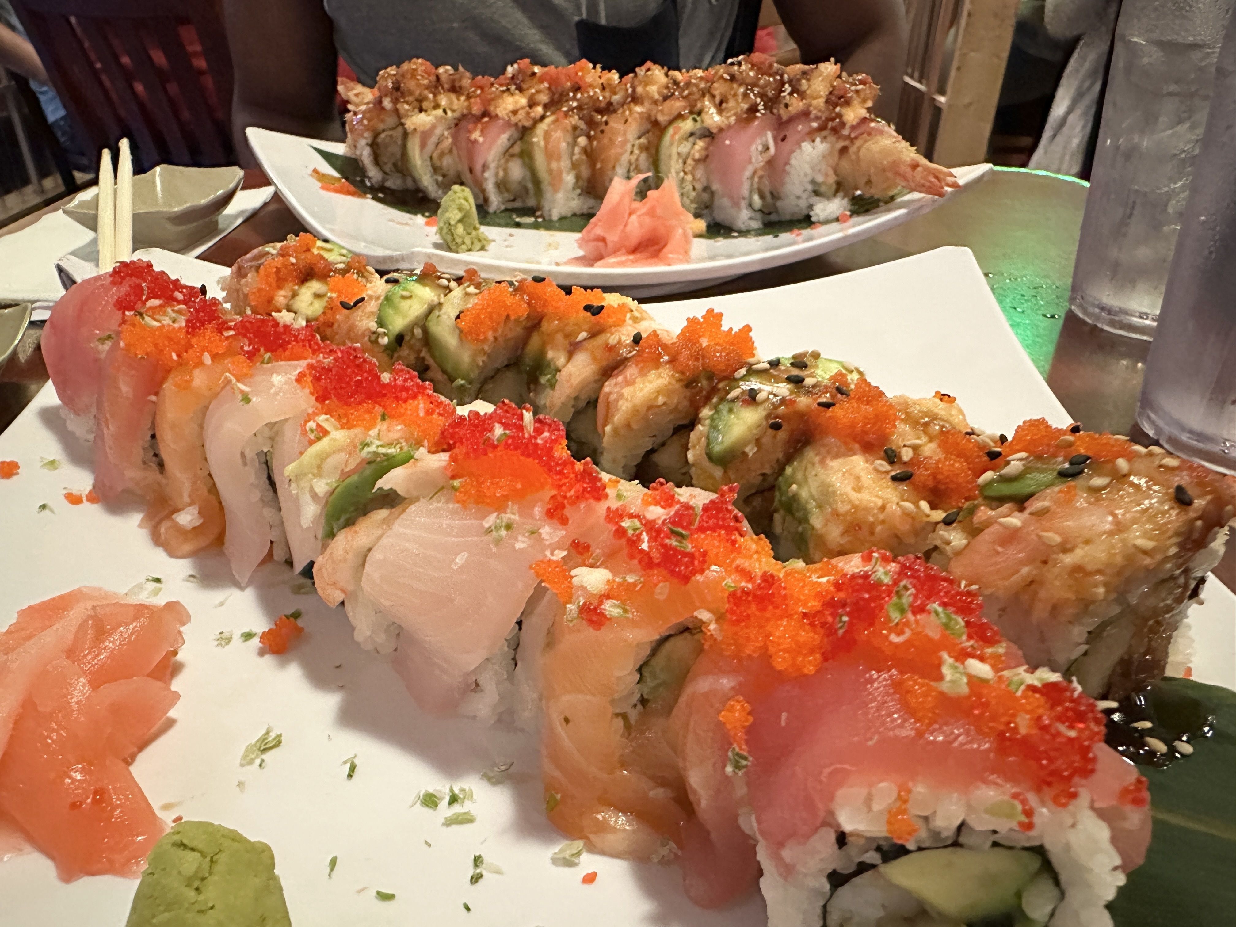 Two sushi plates on a table: foreground shows nigiri topped with orange tobiko and avocado; background displays a long row of crunchy-topped rolls. Wasabi, pickled ginger, chopsticks, and a glass nearby.