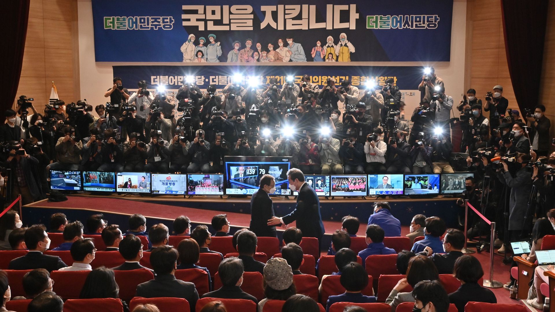 In this image, two men shake hands in front of a stage