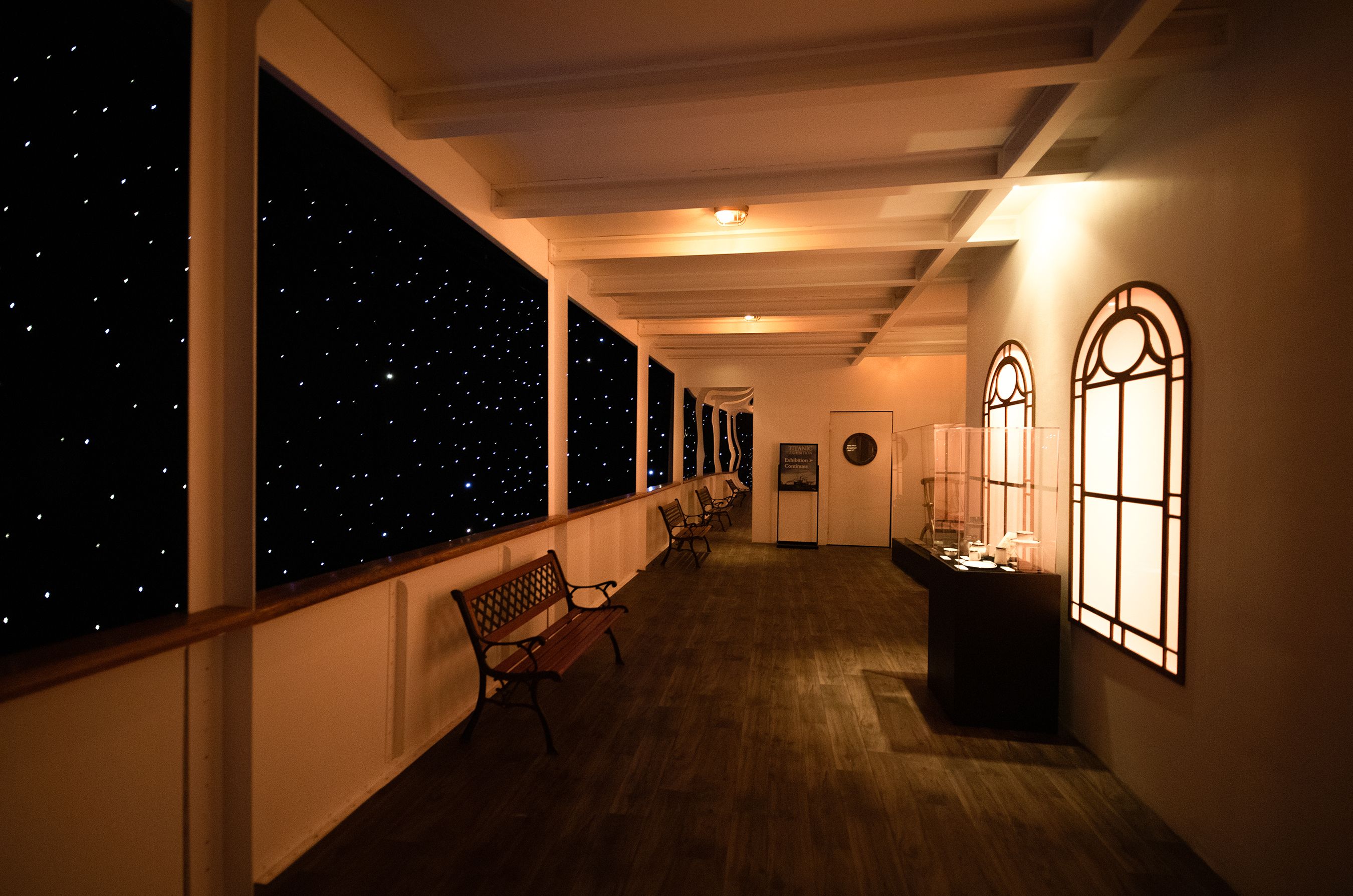 Interior hallway of a museum: a dark, starry wall on the left, white beam ceiling, warm amber lighting, wooden floor, benches along the wall, and illuminated arched display windows on the right.