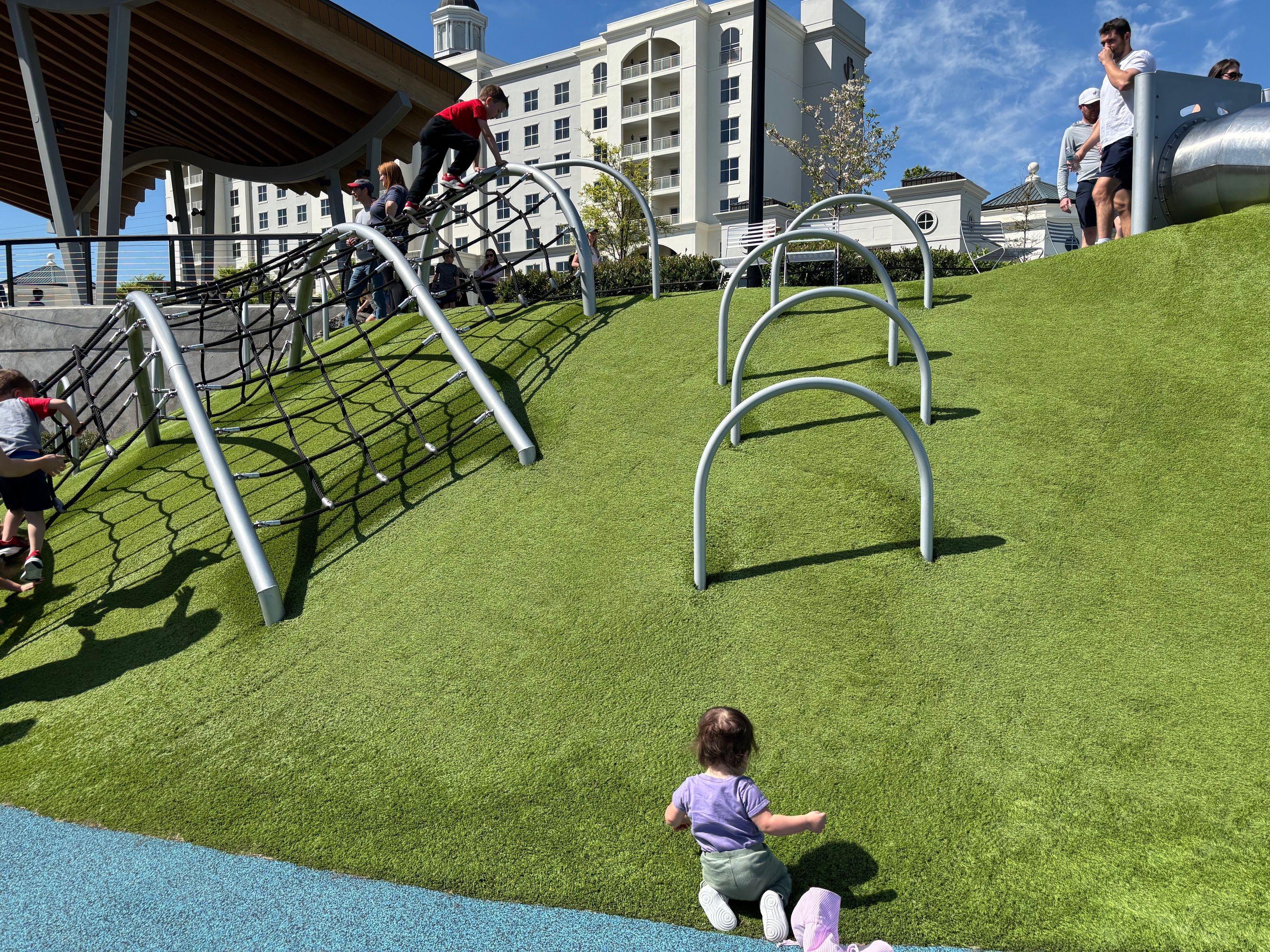 Sunny playground on a grassy hill with a climbing net and curved arches. Children climb while a toddler in purple sits on the grass in the foreground, with a white building in the background.