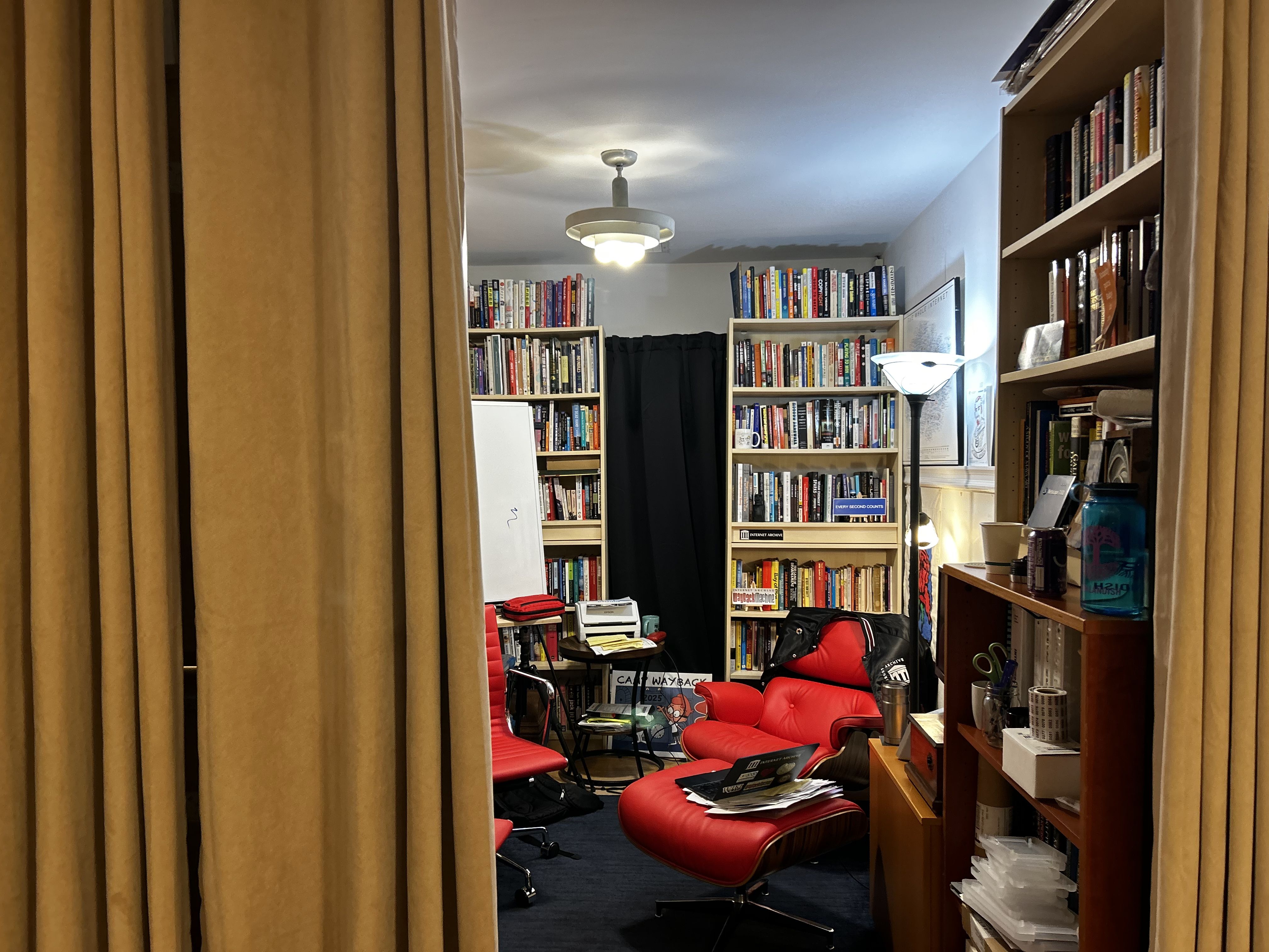 Cozy study room with mustard curtains, red leather chair, bookshelves filled with books, a whiteboard, floor lamp, and various office supplies on shelves and desks.