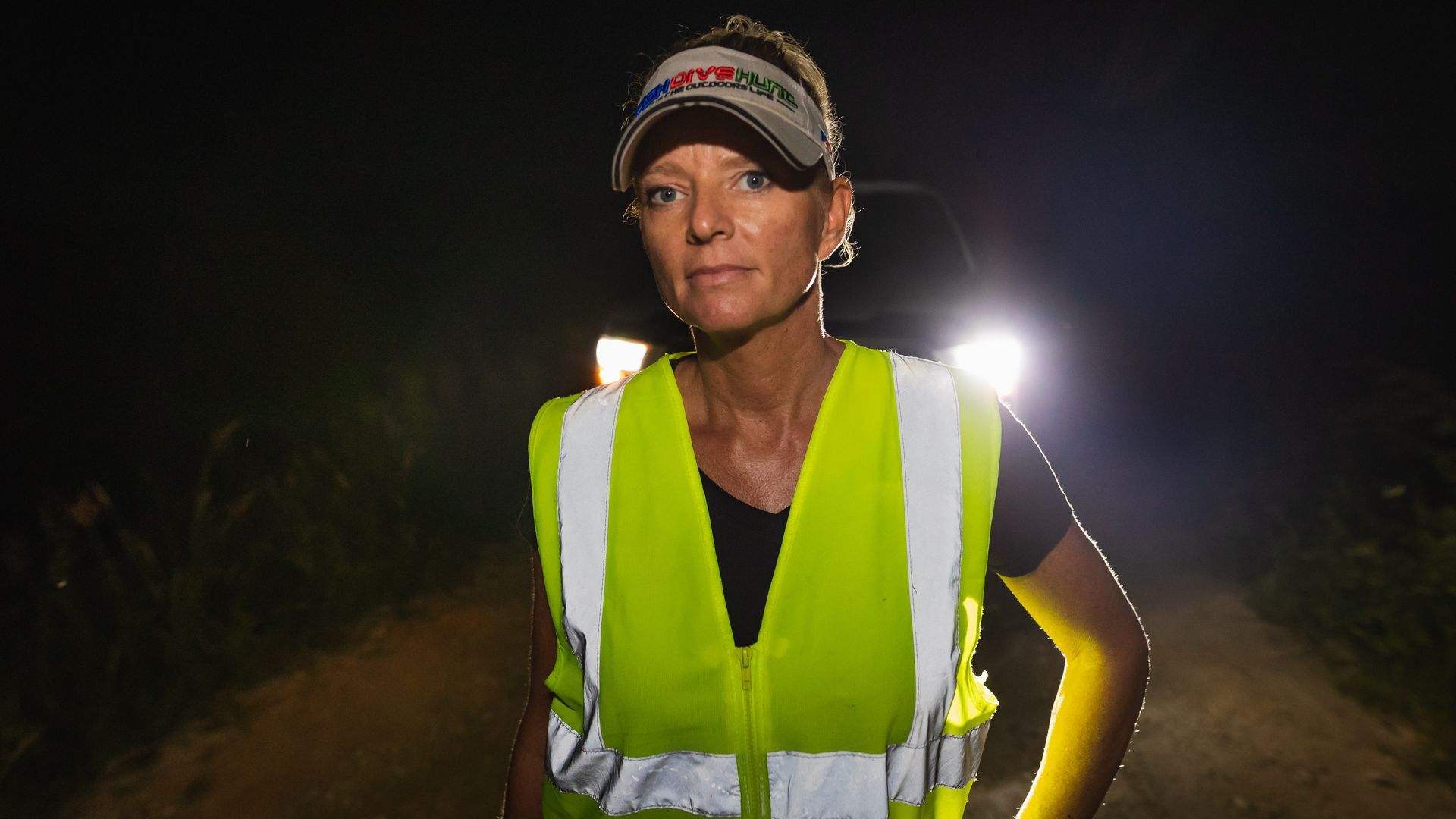 Amy Siewe looks at the camera in reflective gear, backlit by the lights of a pick-up truck.