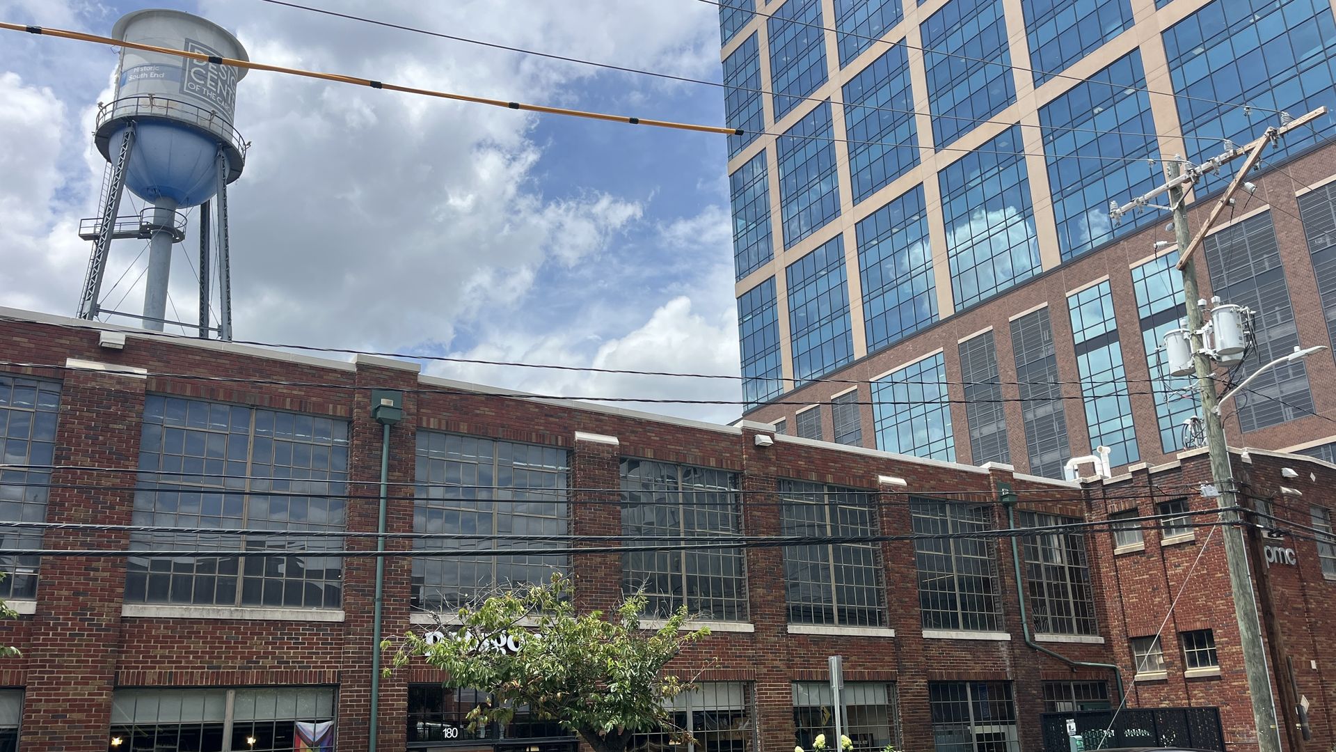 City street with a brick building in the foreground, a blue water tower on the left, a glass office tower on the right, overhead power lines, parked cars, a small tree, and a Pride flag in a storefront window under a blue sky.