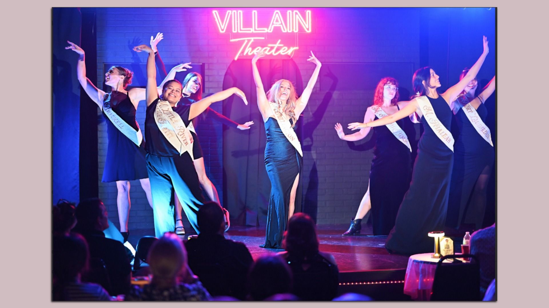 Seven women wearing black dresses or outfits with sashes perform a choreographed dance on stage under a pink neon sign reading "VILLAIN Theater," with an audience watching.
