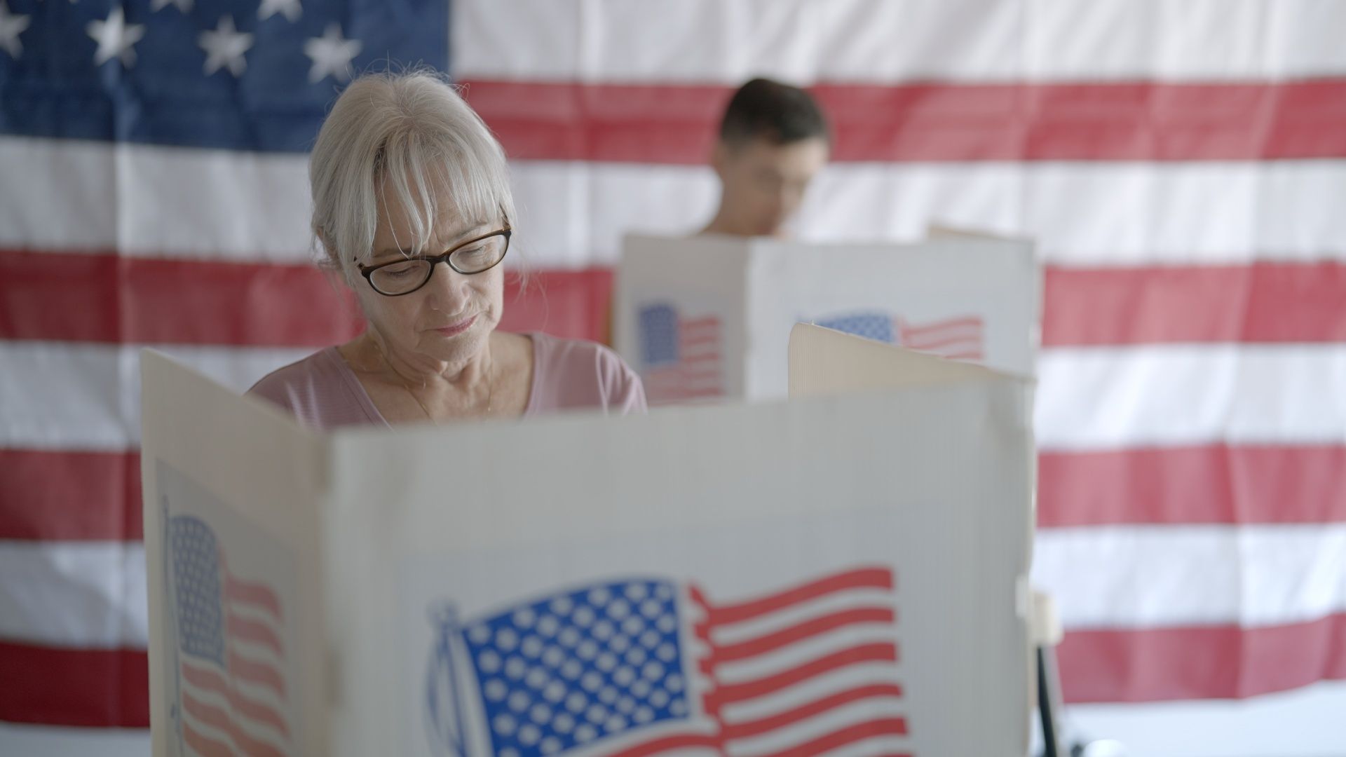 An older woman with gray hair and glasses casts a ballot in a white voting booth, with a large American flag backdrop and a blurred person in the background.