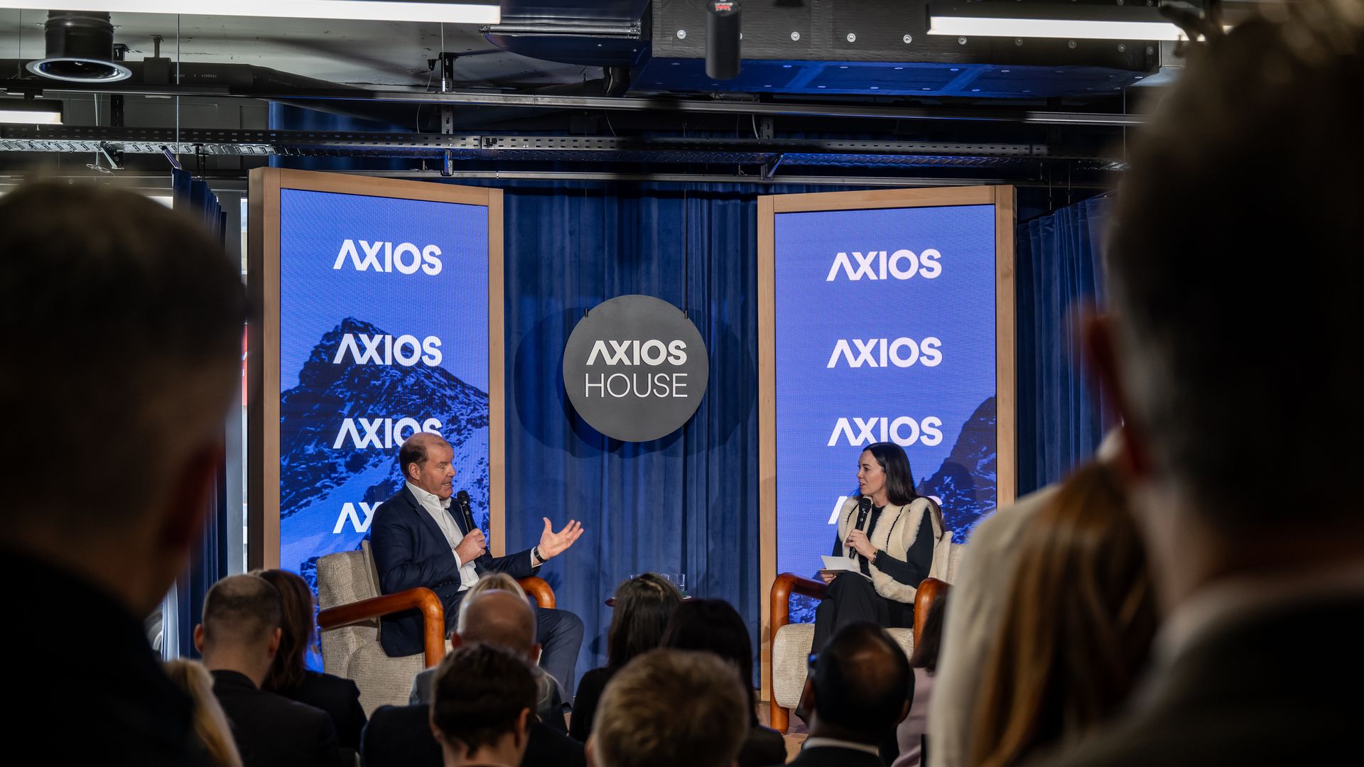 Two people in a discussion on stage at an Axios House event with blue screens displaying "AXIOS" and a black round sign with "AXIOS HOUSE" behind them, audience watching.