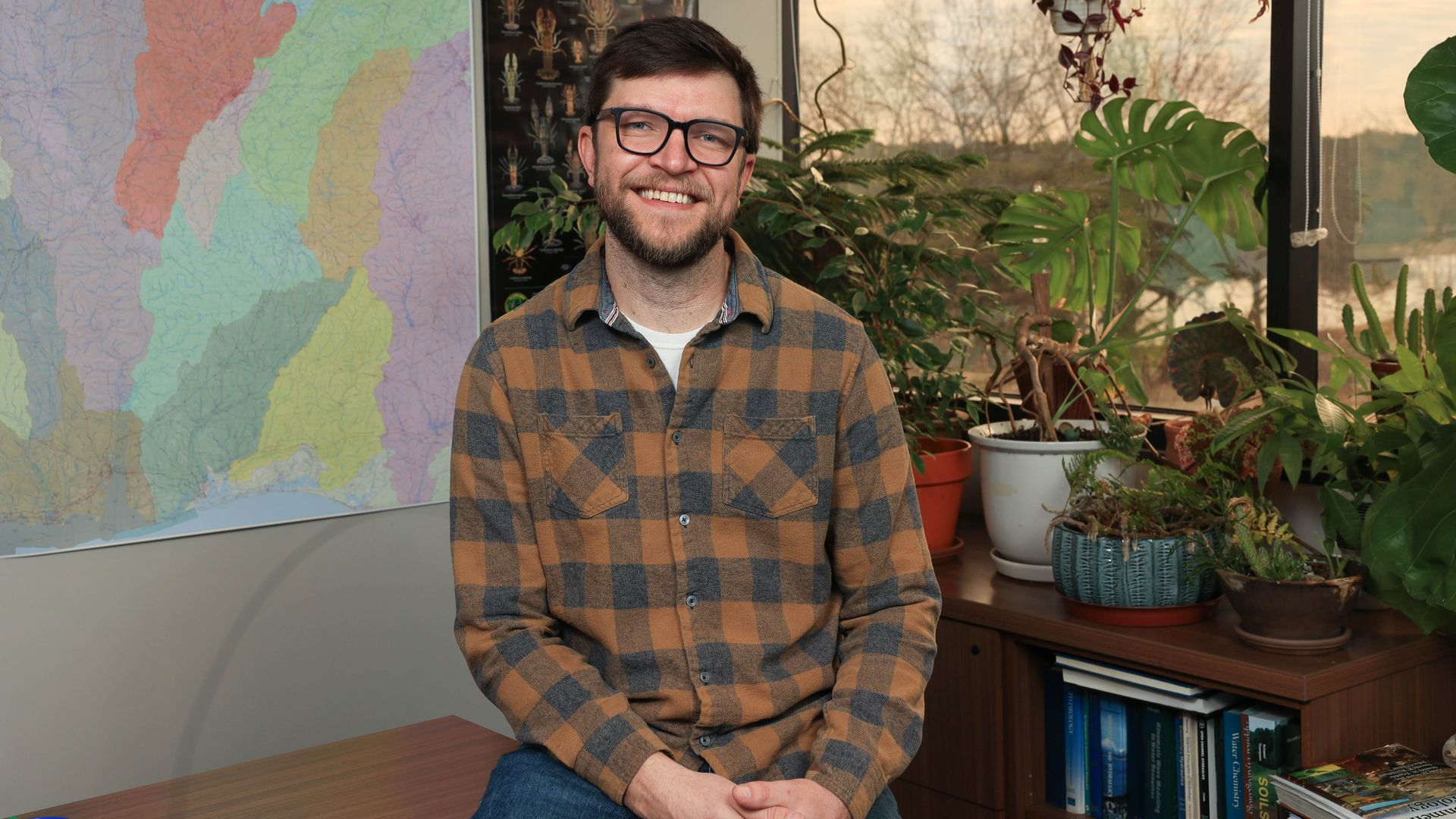 Smiling man with glasses in a brown and black checkered shirt sitting in a plant-filled office with a colorful map and a "Crayfishes of Alabama" poster behind him, sunset outside.