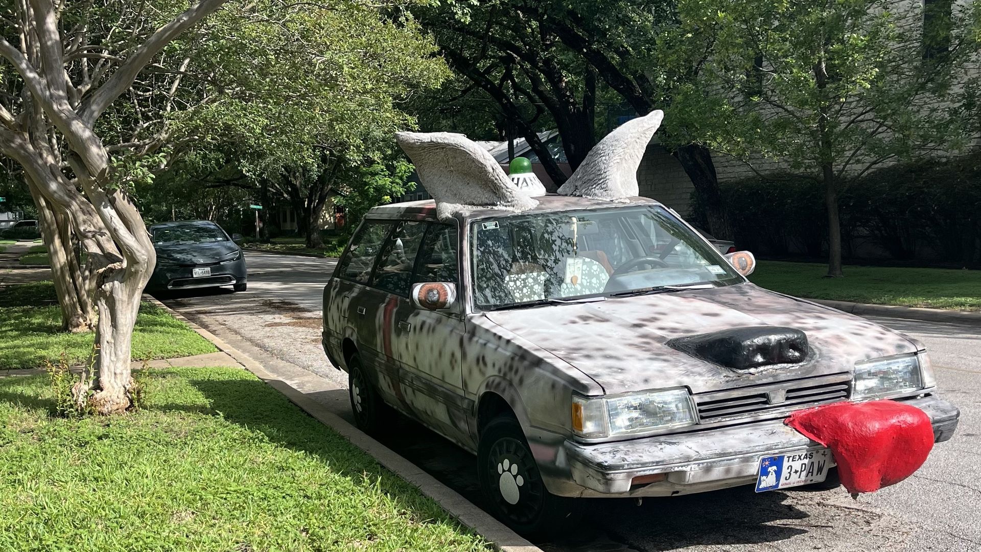 Tree-lined suburban street with a dusty gray SUV topped by two large white foam wing sculptures; a red cloth covers the front bumper and a Texas license plate is visible.
