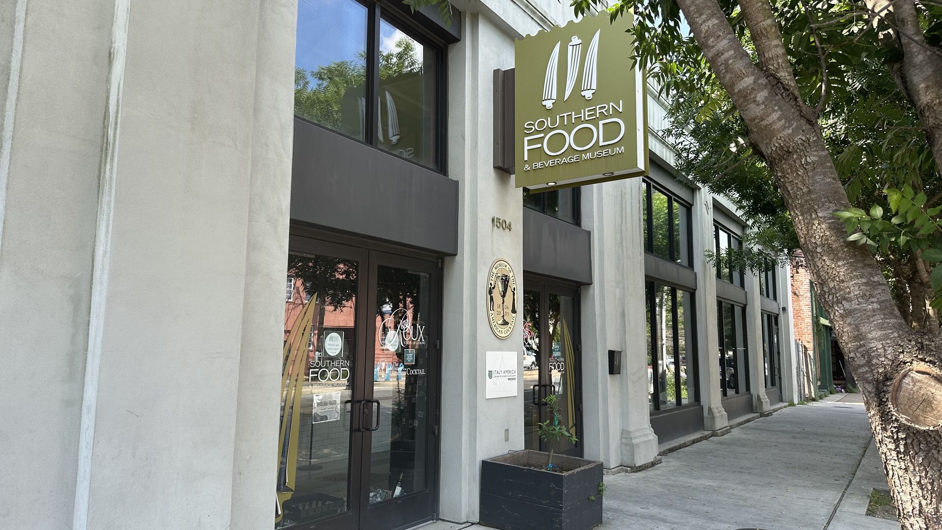 Exterior of a modern gray building with a green sign reading "SOUTHERN FOOD & BEVERAGE MUSEUM" and white utensil graphics; glass doors, sidewalk, and a large tree on the right.