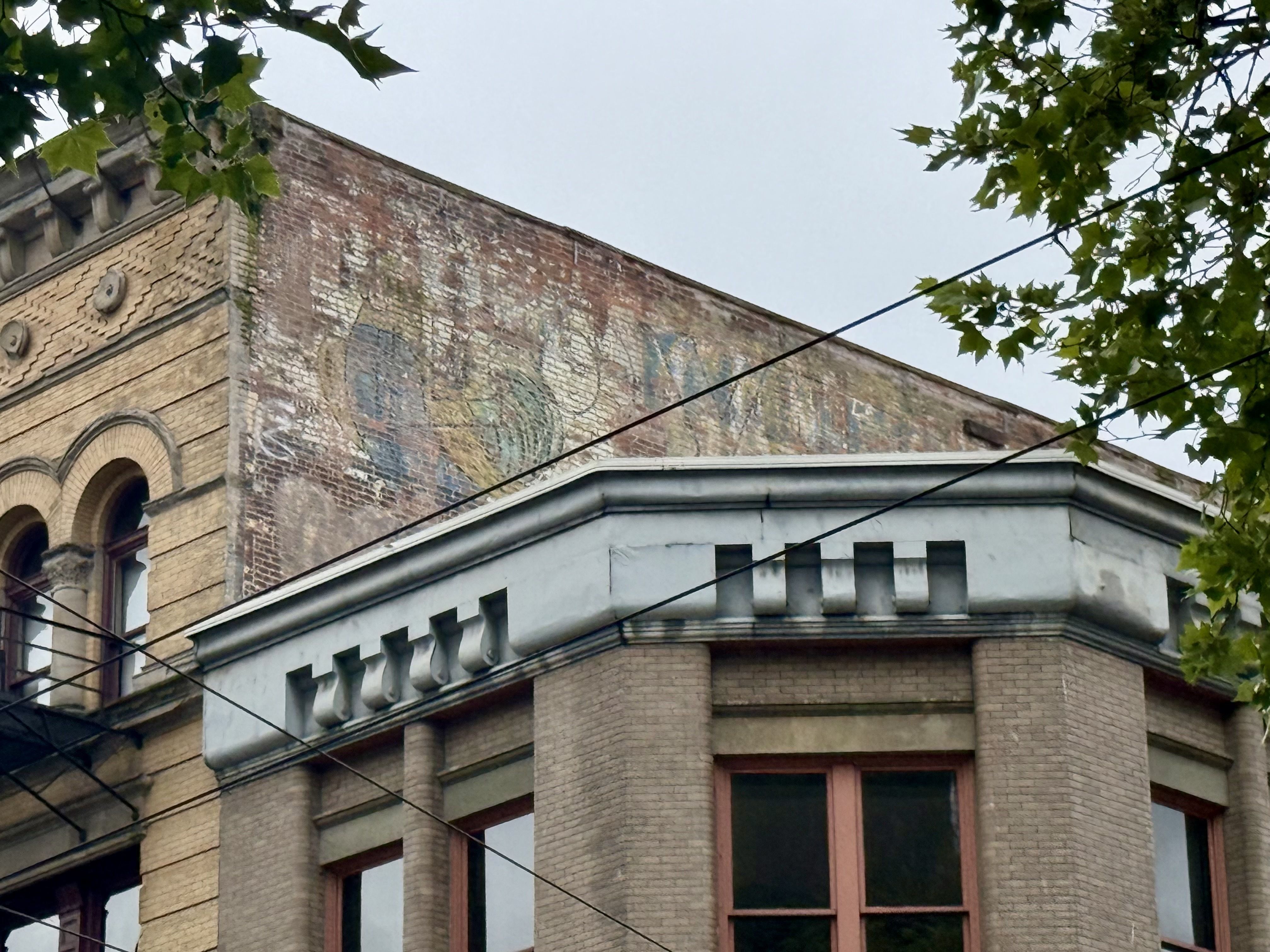 Corner of an old brick building with an arched window and faded mural on the brick wall; decorative cornice, leafy tree branches above, and overhead wires crossing.