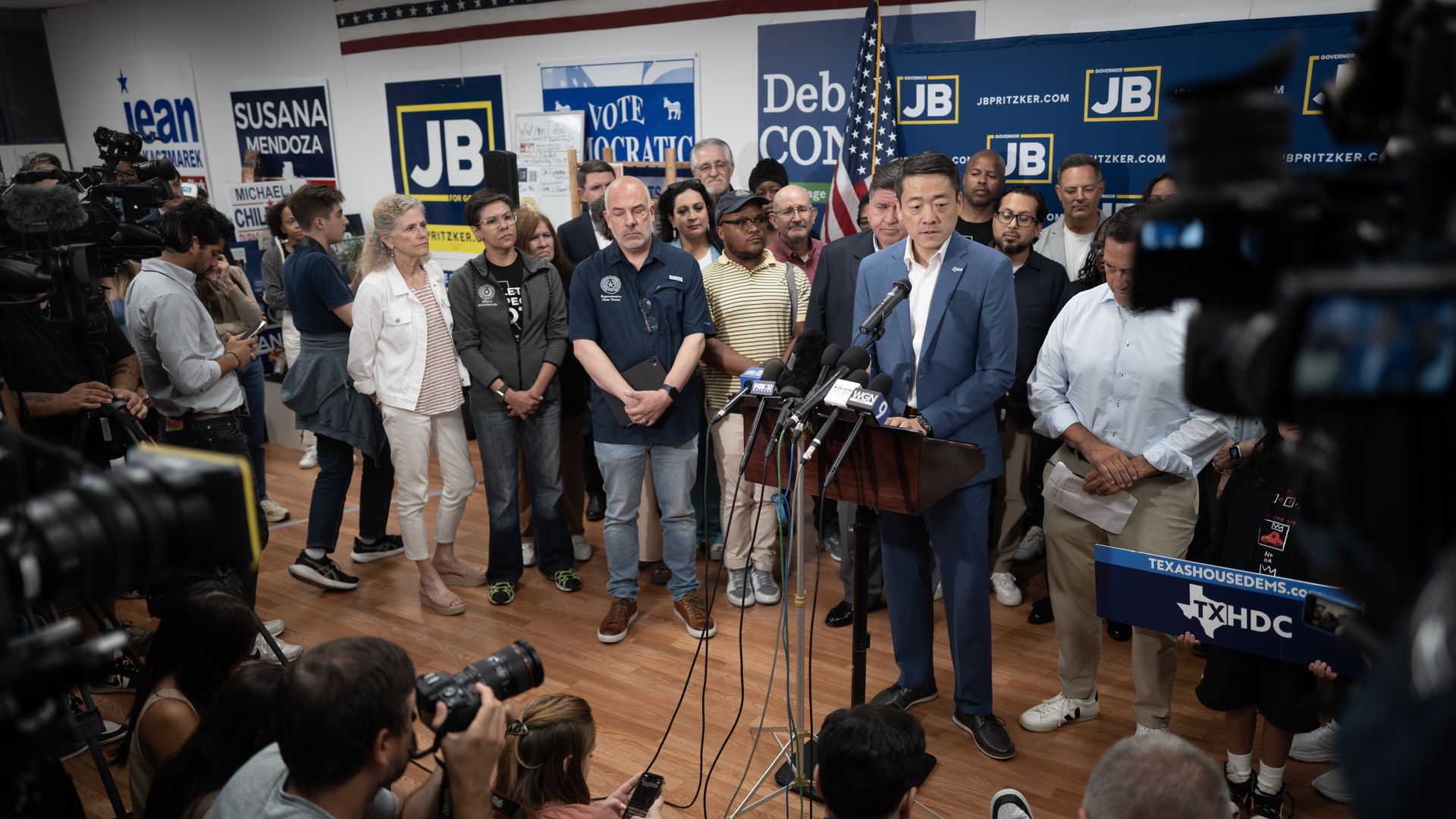Man in blue suit speaks at a podium surrounded by supporters and media with cameras and microphones in a room decorated with political signs and an American flag.