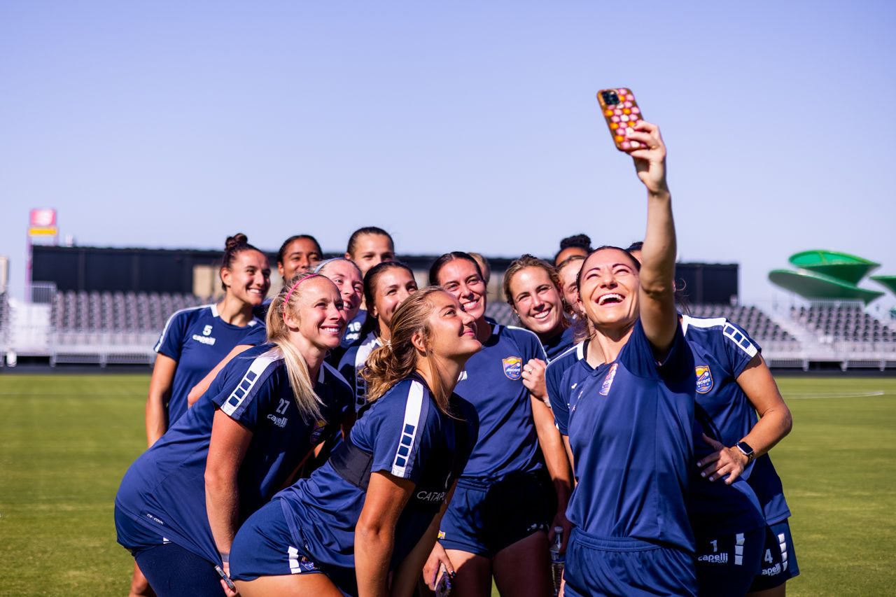 A group of female soccer players in navy blue uniforms with white stripes smiling and posing for a selfie on a sunny soccer field with stands and a clear blue sky in the background.