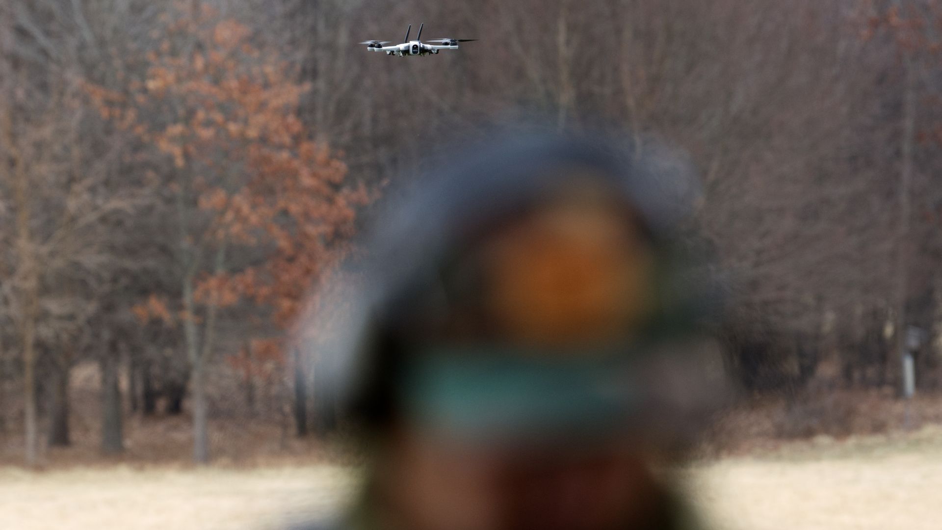 A small drone hovers behind a man, whose face is in the foreground and is very out of focus. The background is field and trees. It looks like winter without the snow.