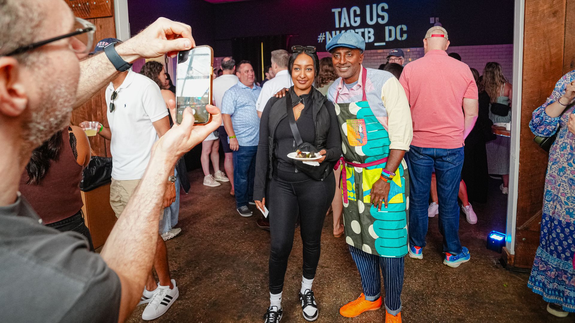 A man in a bright patterned outfit and a woman in black pose in a doorway at a crowded party; someone in the foreground holds a smartphone, with a neon "TAG US" sign in the background.