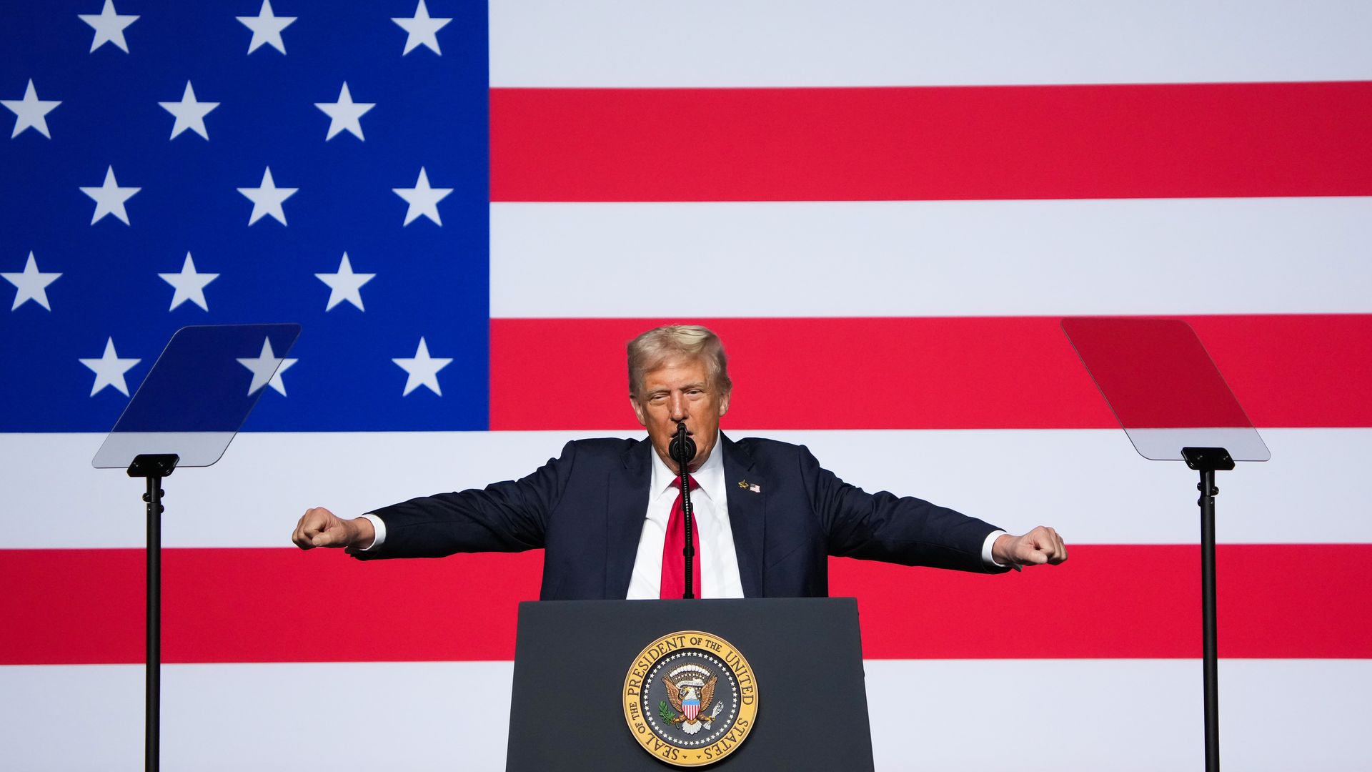 Trump holds his arms out as he speaks from behind a podium in front of a large American flag.