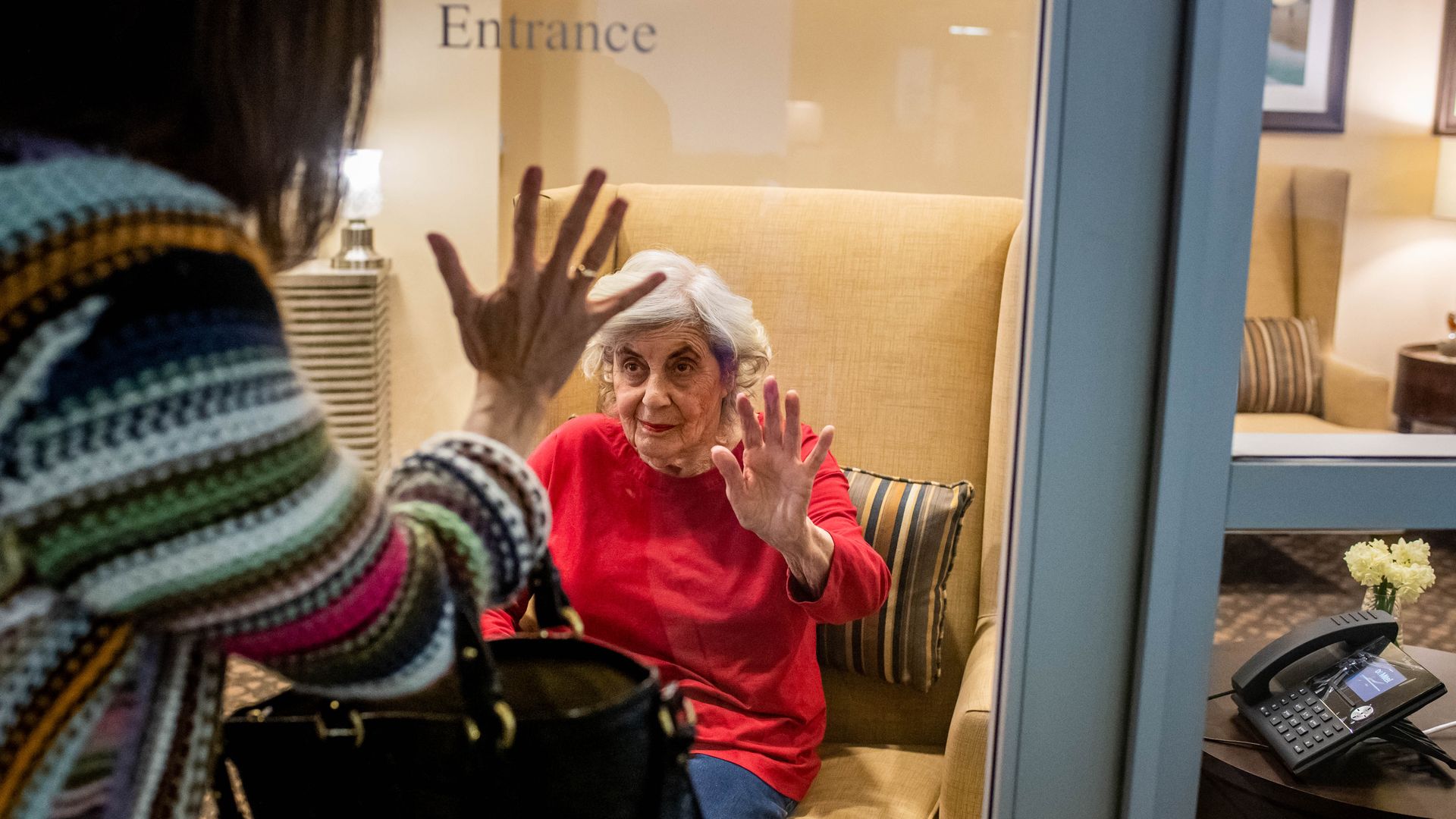 Photo of a nursing home resident holding her hand up to the clear glass as on the other side her daughter raises her own hand