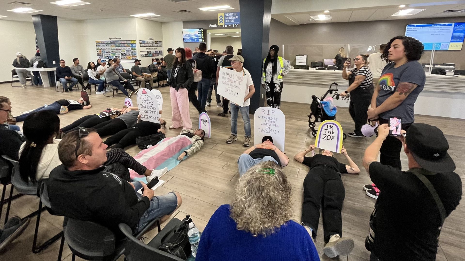 Several people lie on the ground of a government office holding signs shaped like tombstones. "RIP Attacked by Florida Legislature and DHSMV," one says. "Remember me," another says, held by a person who is covered by a blue, pink and white transgender flag. Onlookers stare and take photos.