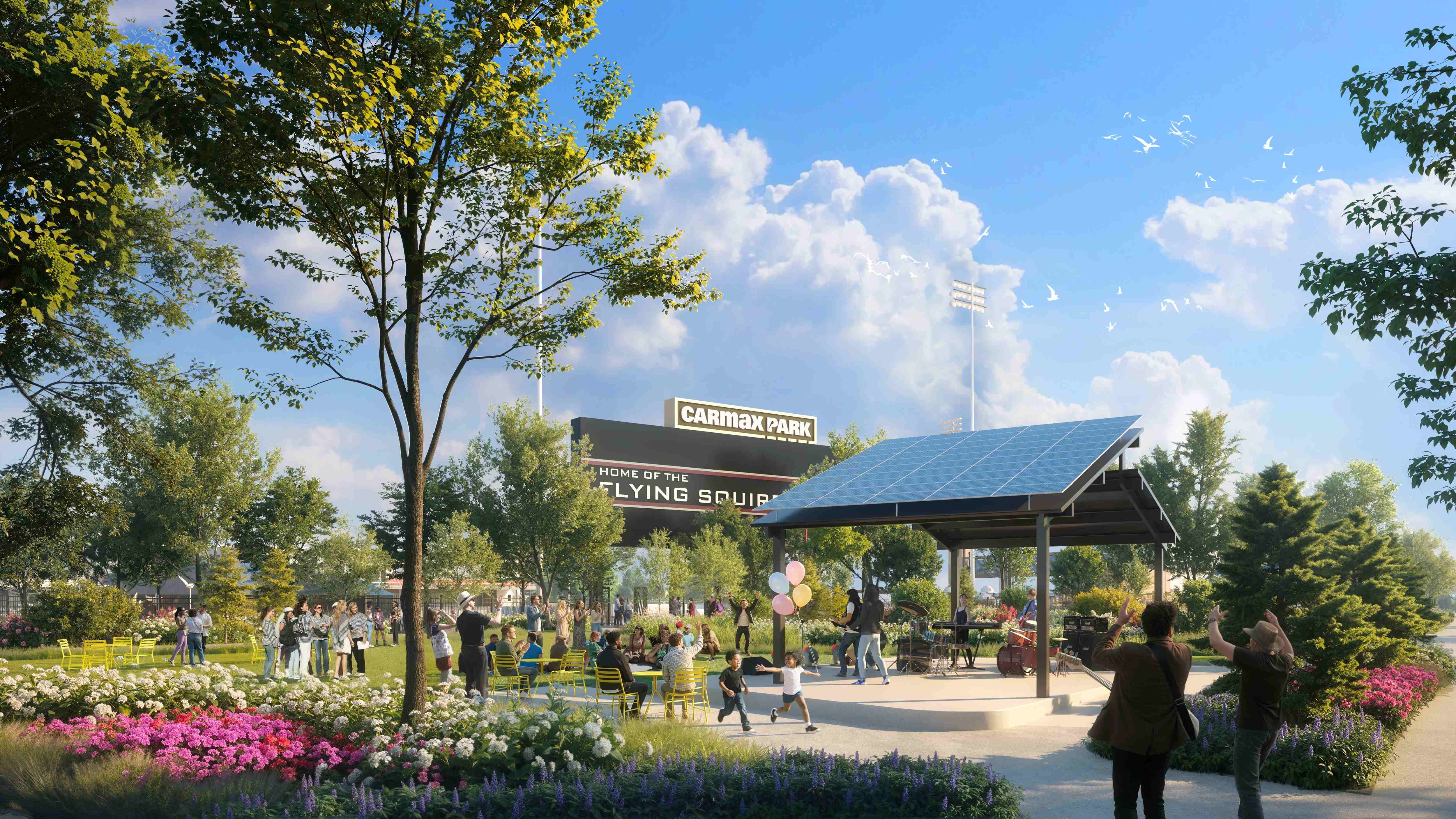 Sunny park scene with families, colorful flowers, and trees. A covered pavilion, solar panels, balloons, and a stadium sign reading "CarMax Park" with "Flying Squirrels" nearby.