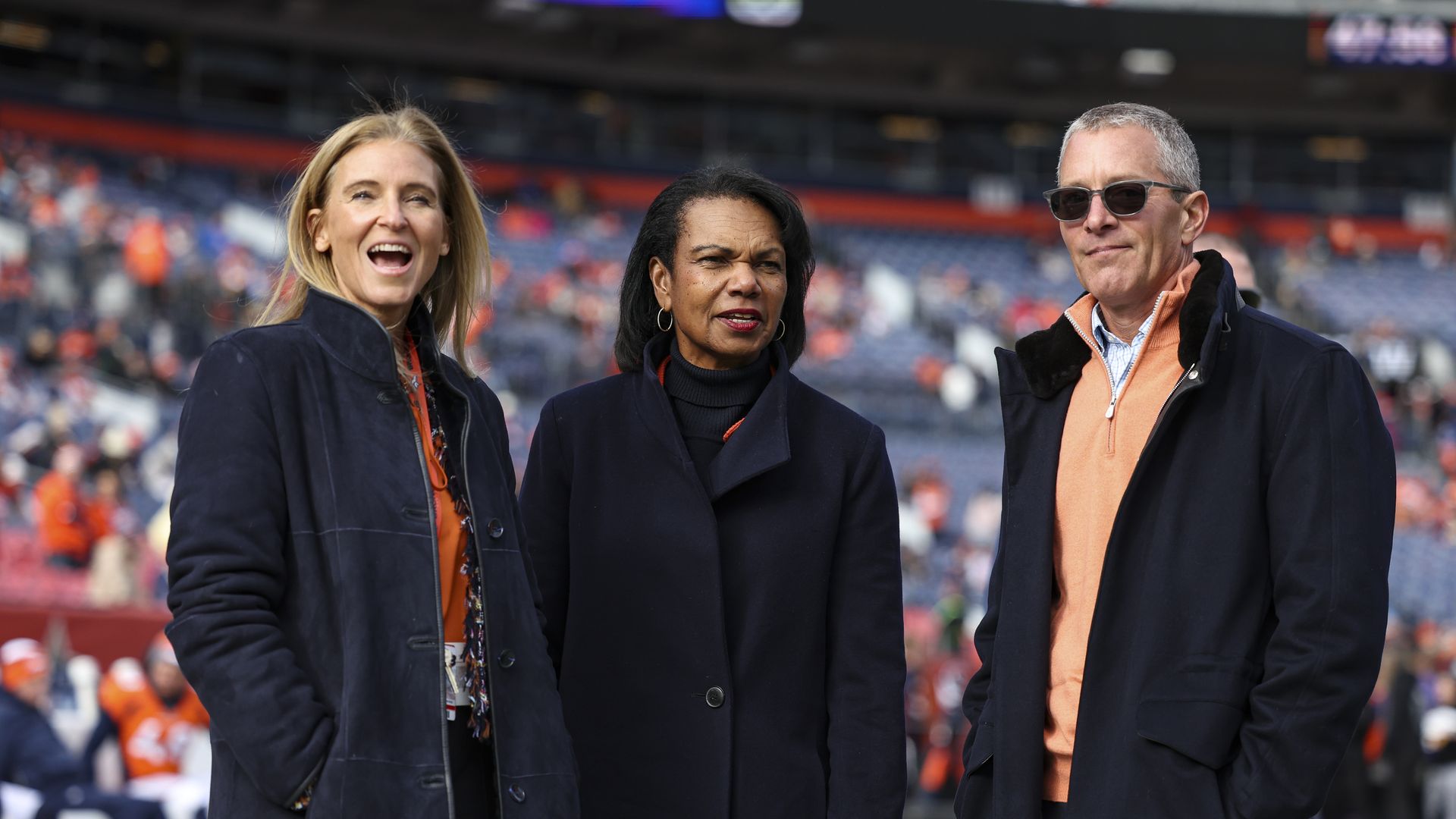 Three people in heavy jackets stand inside a stadium while looking on. 