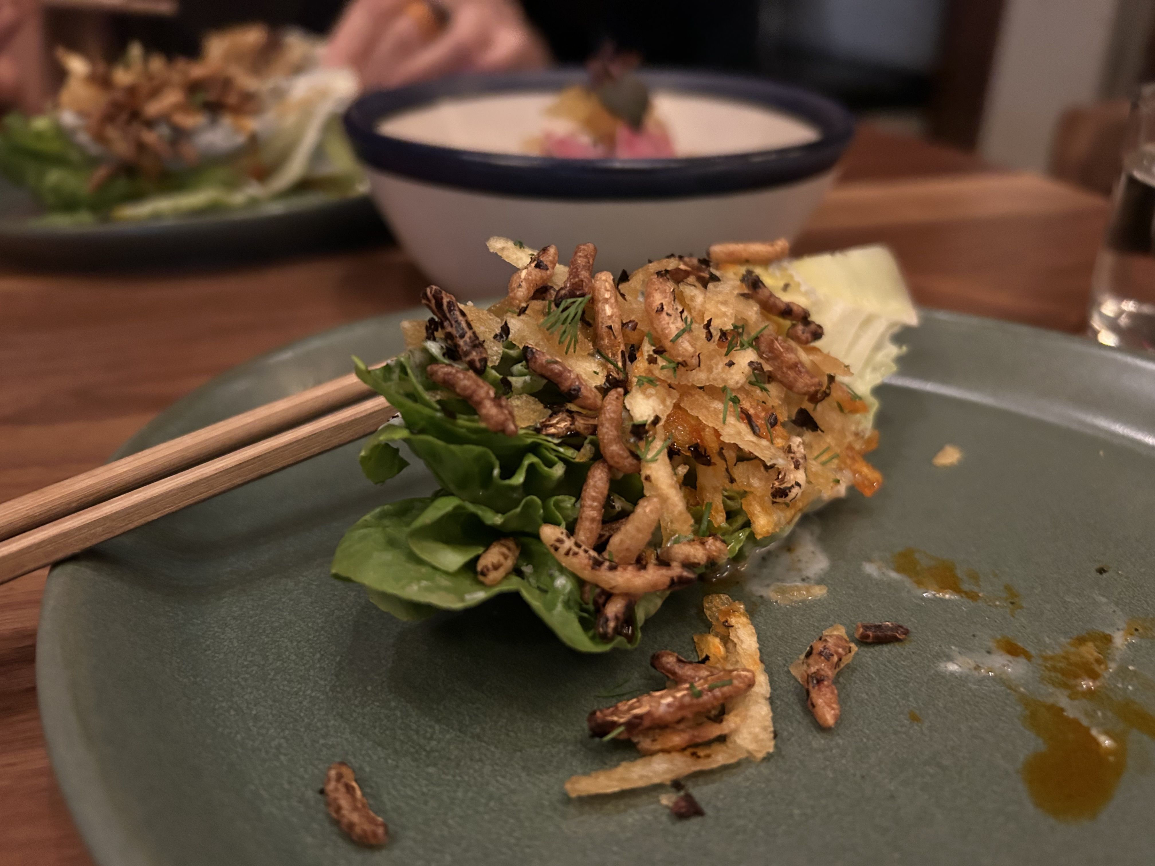 A green leaf lettuce topped with crispy garnishes and herbs on a green plate, with chopsticks resting on the plate and a bowl with food blurred in the background.