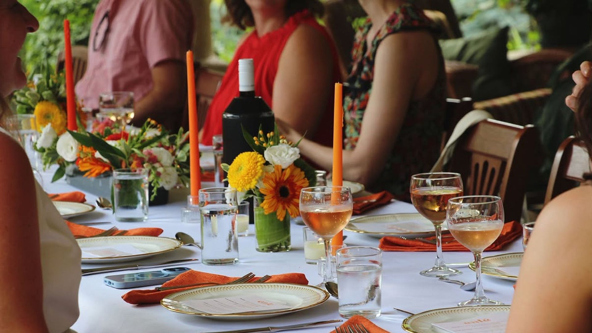 Outdoor dining table set for a celebration with orange candles, yellow and orange flowers, a wine bottle, glasses, gold-rimmed plates, and orange napkins; guests chat in summer dresses.