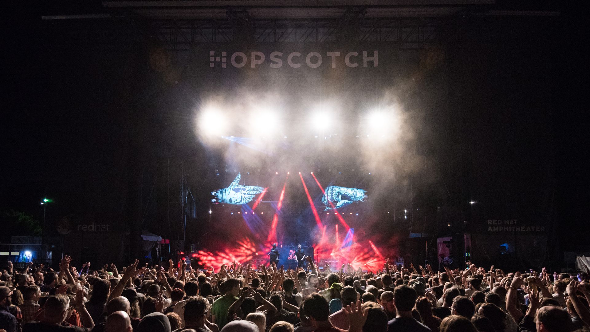 Large crowd at a nighttime concert with bright stage lights, smoke effects, and two large blue hand images on screen under the "OPSCOTCH" sign at Red Hat Amphitheater.