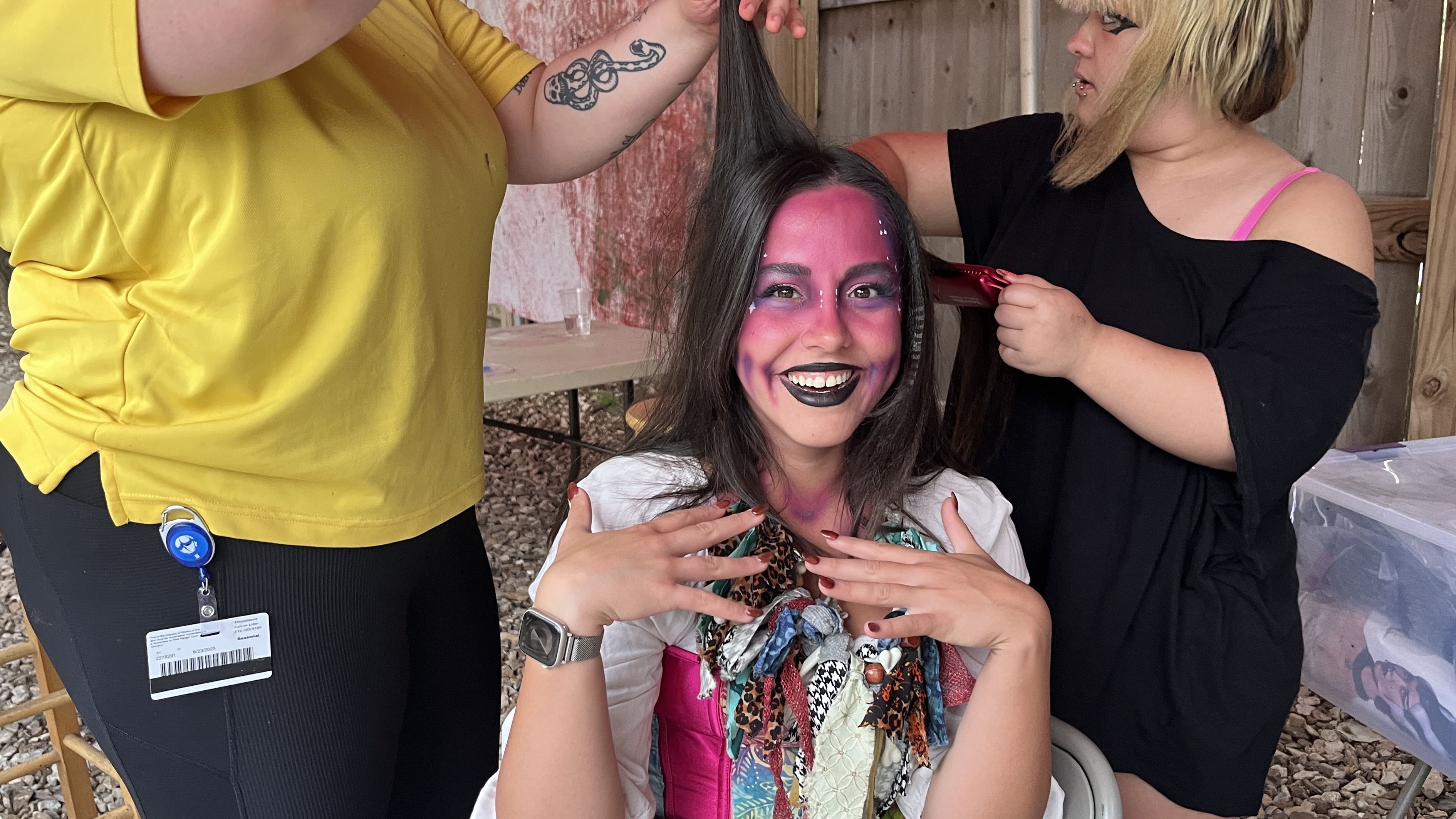 Person with purple face paint, black lipstick, and colorful scarf sits smiling as two others style their long black hair indoors with wooden walls.