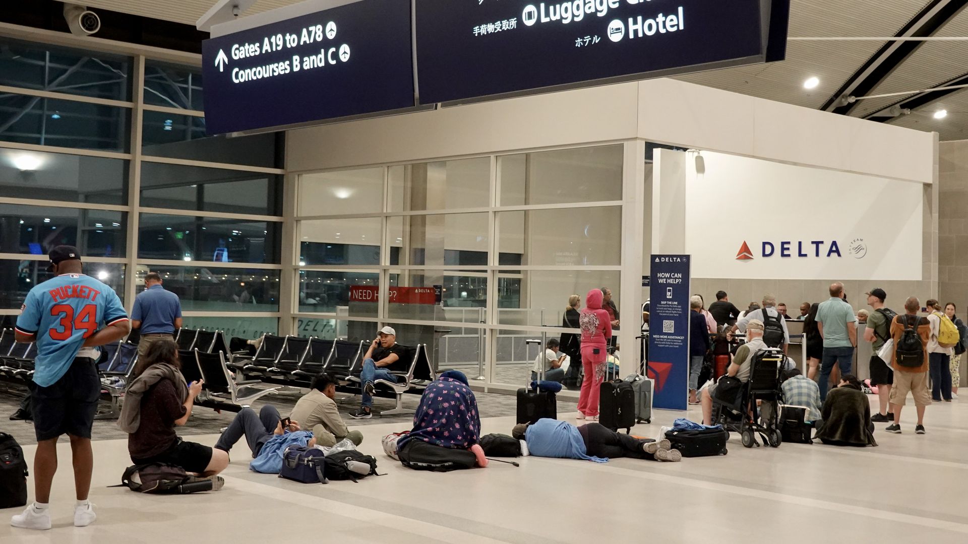 Passengers lay down, sit and stand waiting in line near a Delta sign 