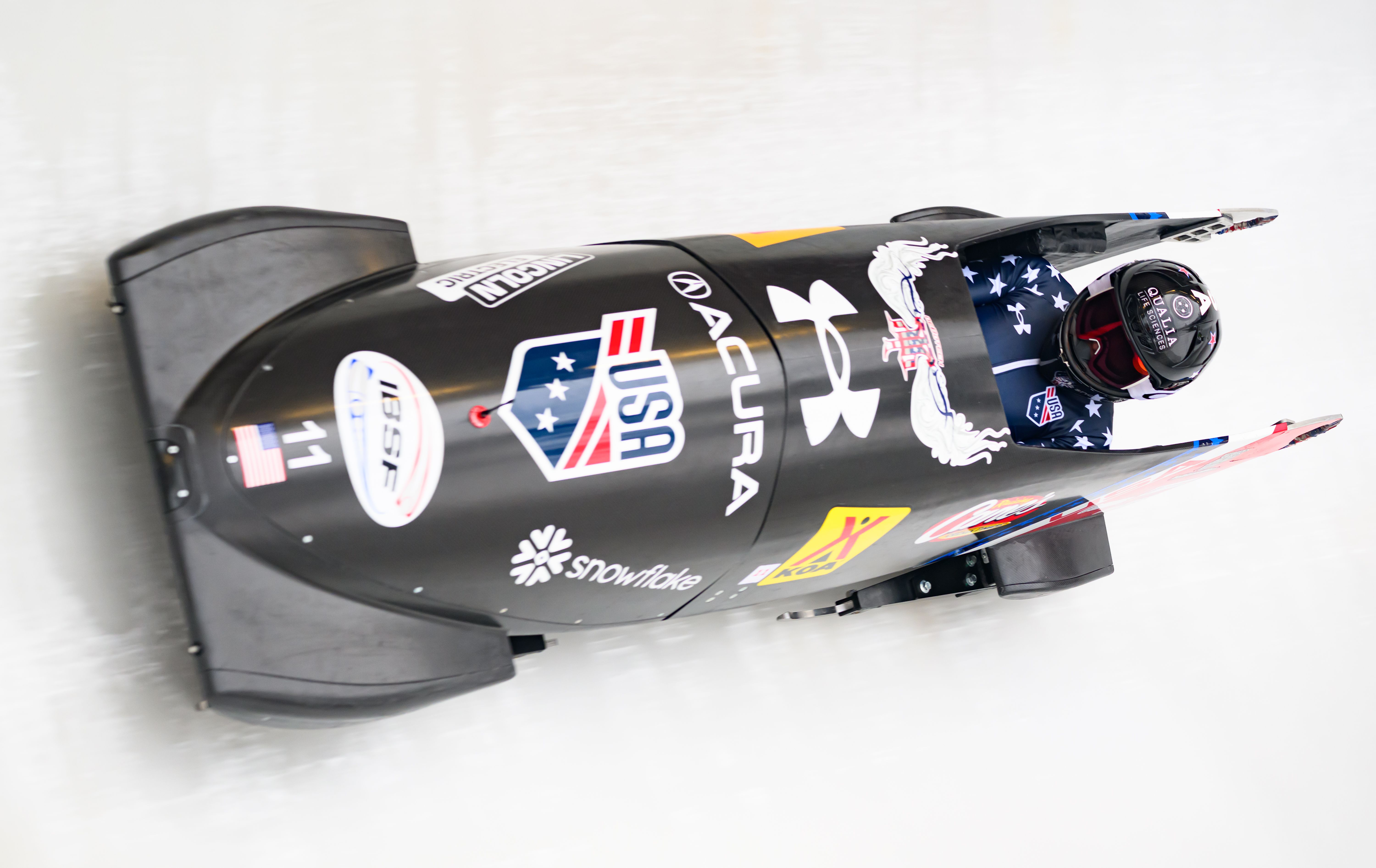 An Olympic athlete with a black helmet and blue suit with white stars rides in a black Team USA branded bobsled with sponsor stickers during a run on the icy track.