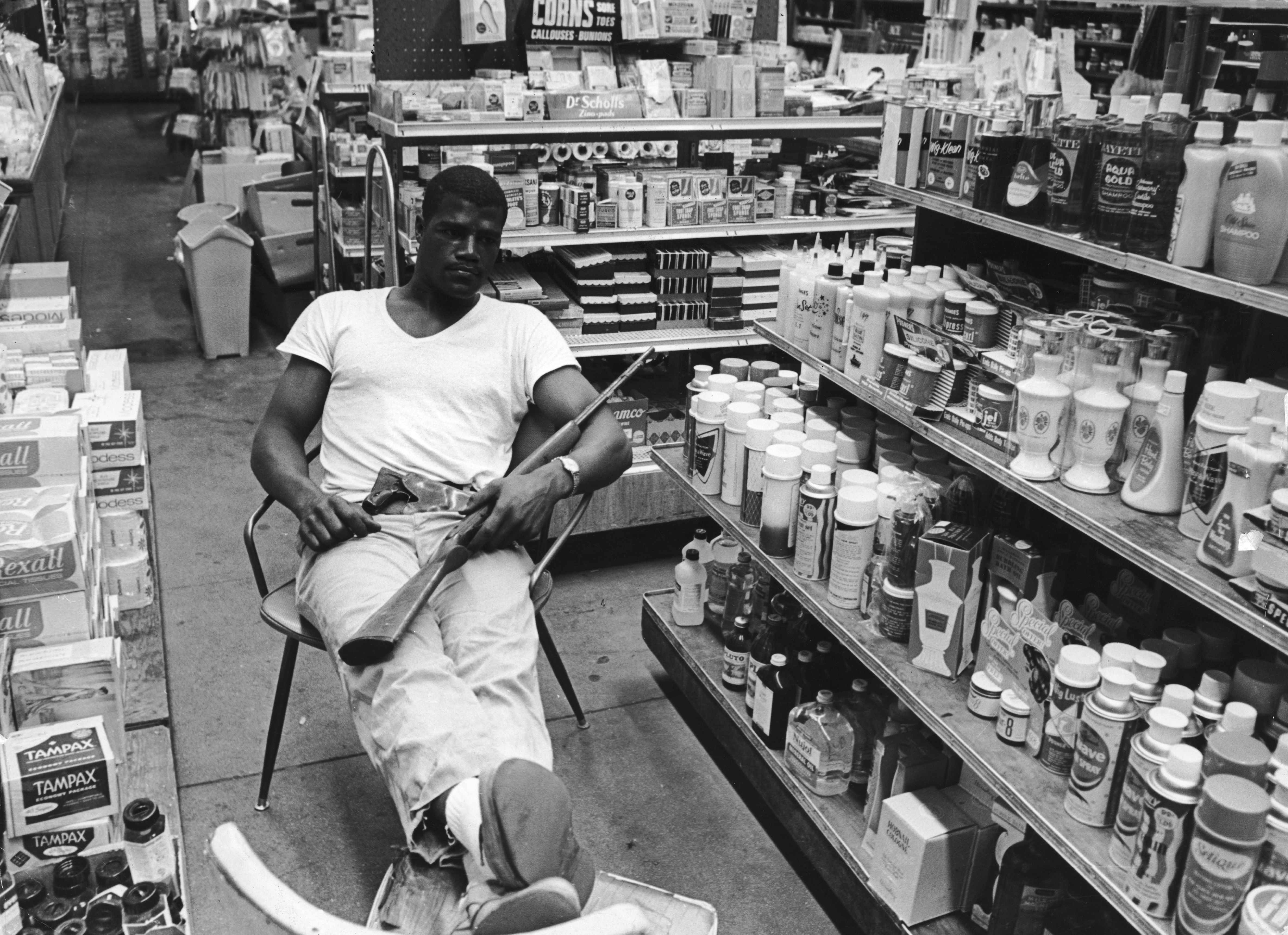 Black and white photo of a man in a white t-shirt and light pants sitting in a chair inside a store, holding a shotgun, surrounded by shelves stocked with various bottles and boxes.