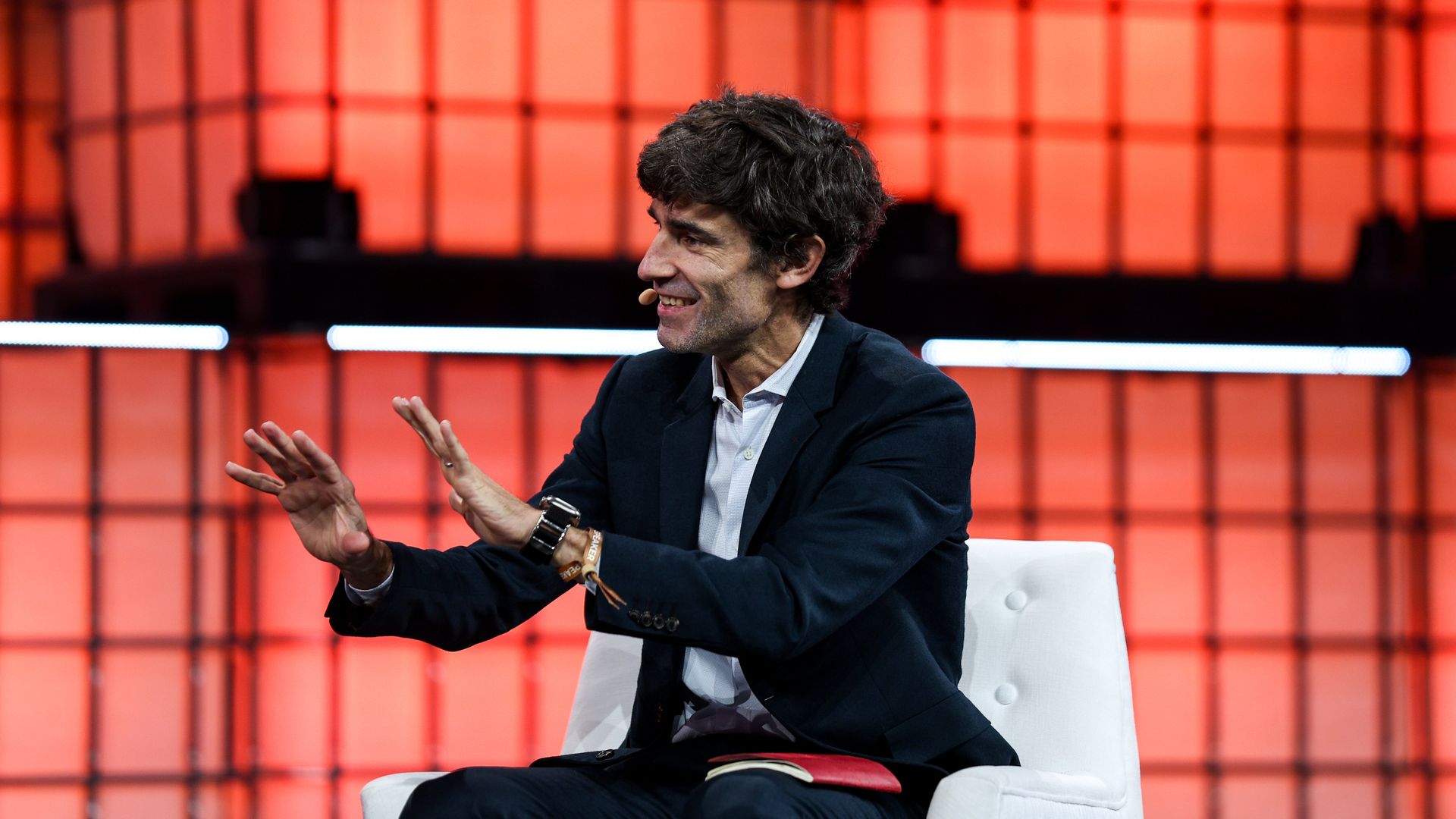 Atlantic CEO Nick Thompson speaks on stage during a conference, gesturing with his hands while seated in a white chair against an orange-lit backdrop.