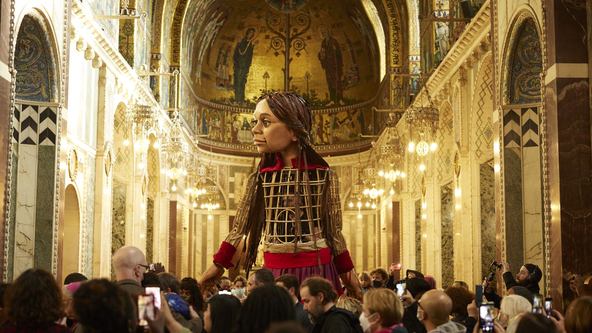 Little Amal, a 12-foot puppet, towers over a crowd of people taking photos inside a cathedral. 
