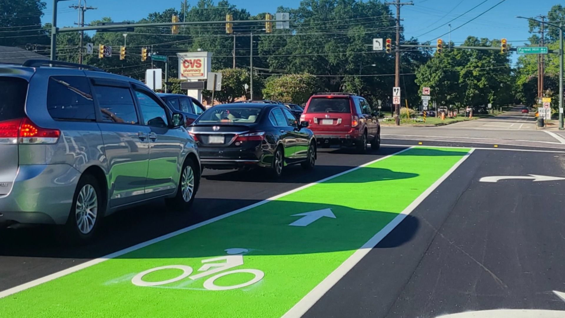 Busy urban intersection with a vivid green bike lane in the foreground, white bike symbol and arrow. Cars line up, CVS Pharmacy sign in the background, traffic lights and trees overhead.
