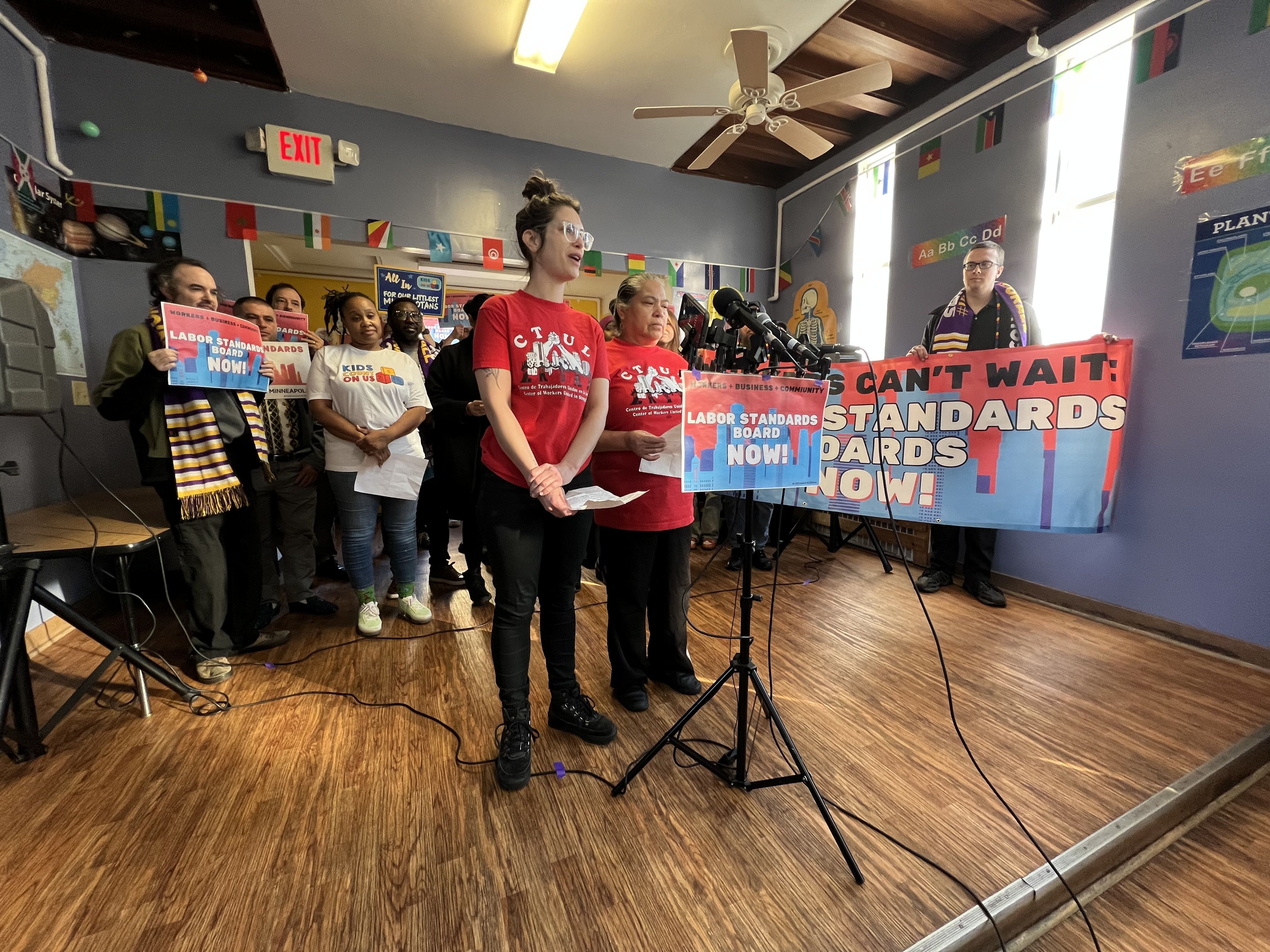 People in red shirts speak at a microphone stand during a press conference in front of a banner reading "Labor Standards Boards Now!"