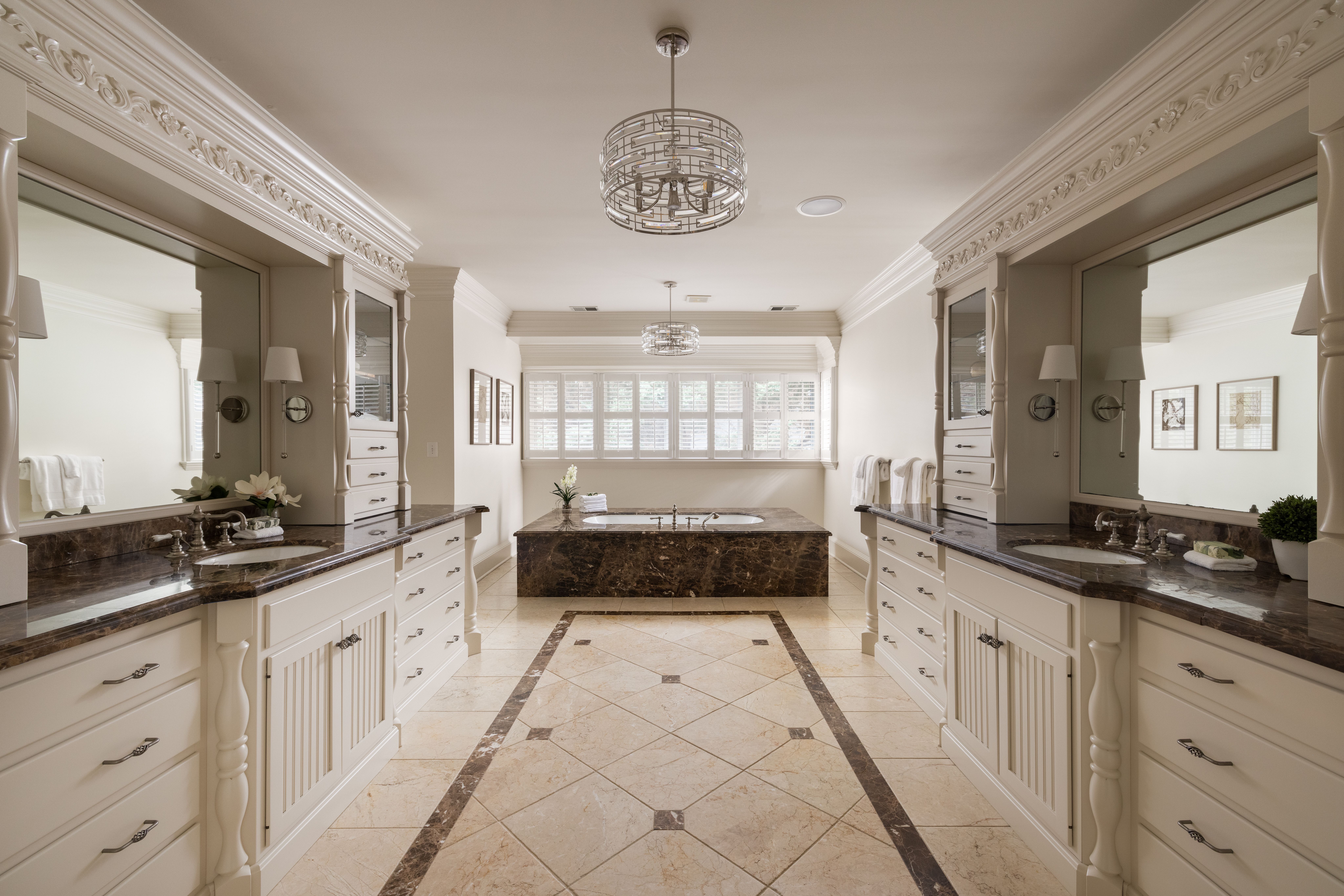 Luxurious bathroom with white cabinetry, dark marble countertops, side vanities, ornate crown molding, a central island, crystal chandelier, and large back windows with shutters.