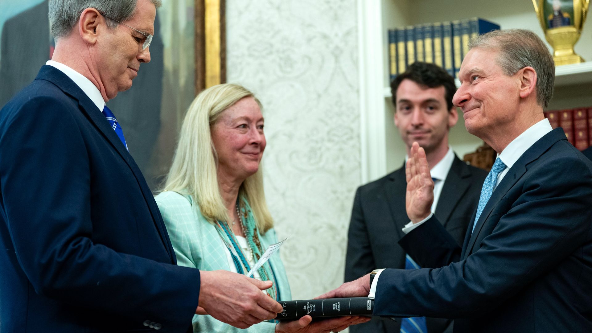 A man holds up his hand with another hand on what appears to be a Bible, held by another man. A woman and a young man look on, inside what appears to be an ornate office, with lots of gold. 