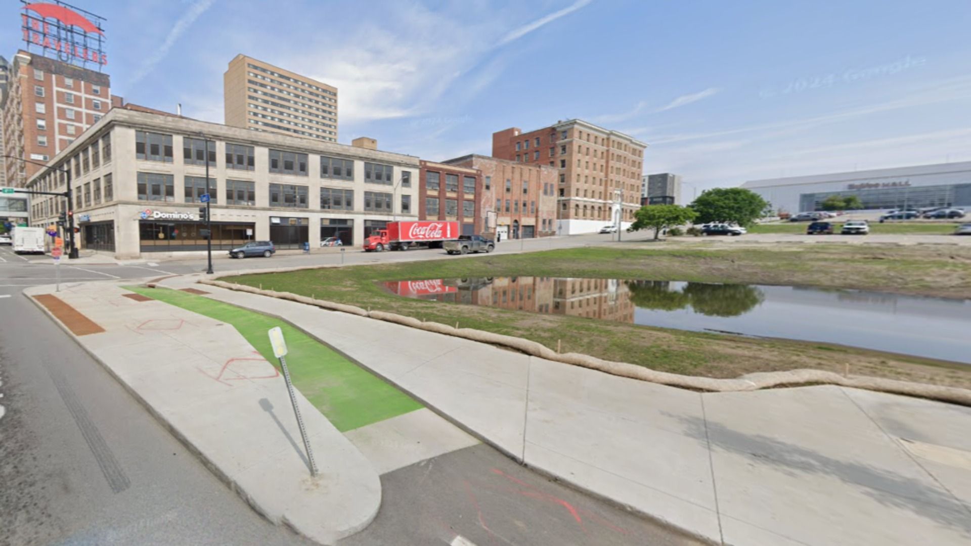 Street view of a downtown block with a Domino's on the left, brick buildings, a red Coca-Cola truck, a reflective canal in the foreground, and a large mall in the distance under a blue sky.
