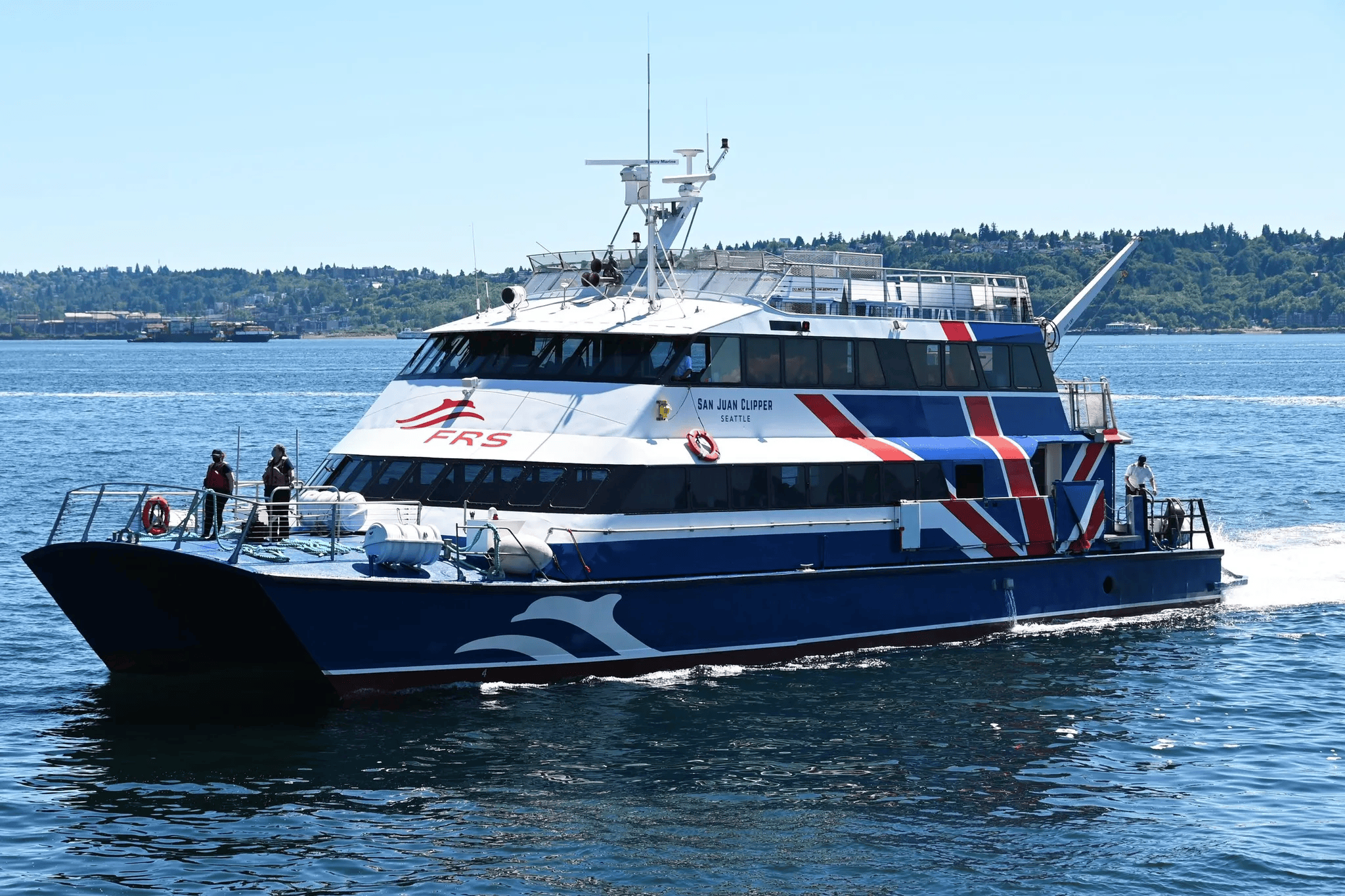 White and blue passenger ferry with red accents glides on calm water; hull reads "San Juan Clipper, Seattle." A tall radar mast and deck gear rise above, with a forested shoreline in the distance.