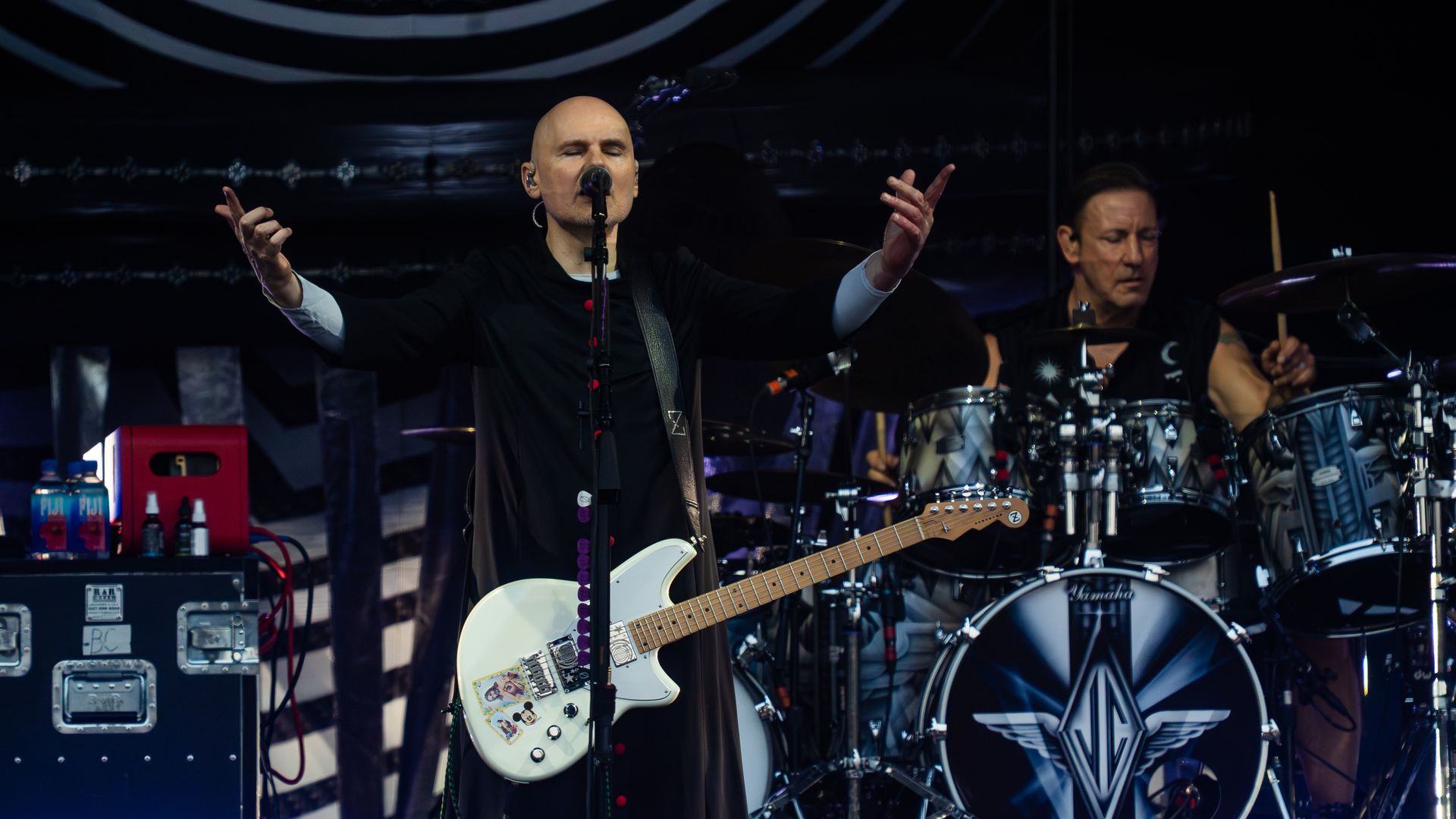Bald male singer with a white electric guitar, stickers on the body, arms raised, singing into a microphone as a drummer plays behind on a dark stage.
