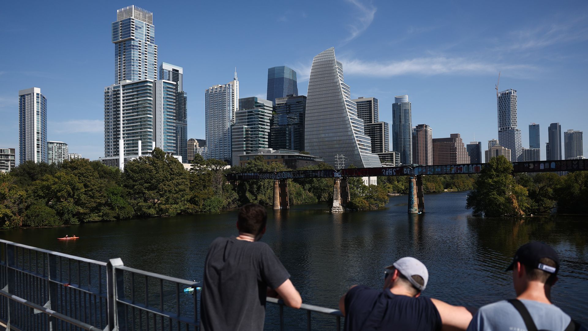 Three people stand on a bridge overlooking a river with a city skyline of modern high-rise buildings and trees in the background under a clear blue sky.