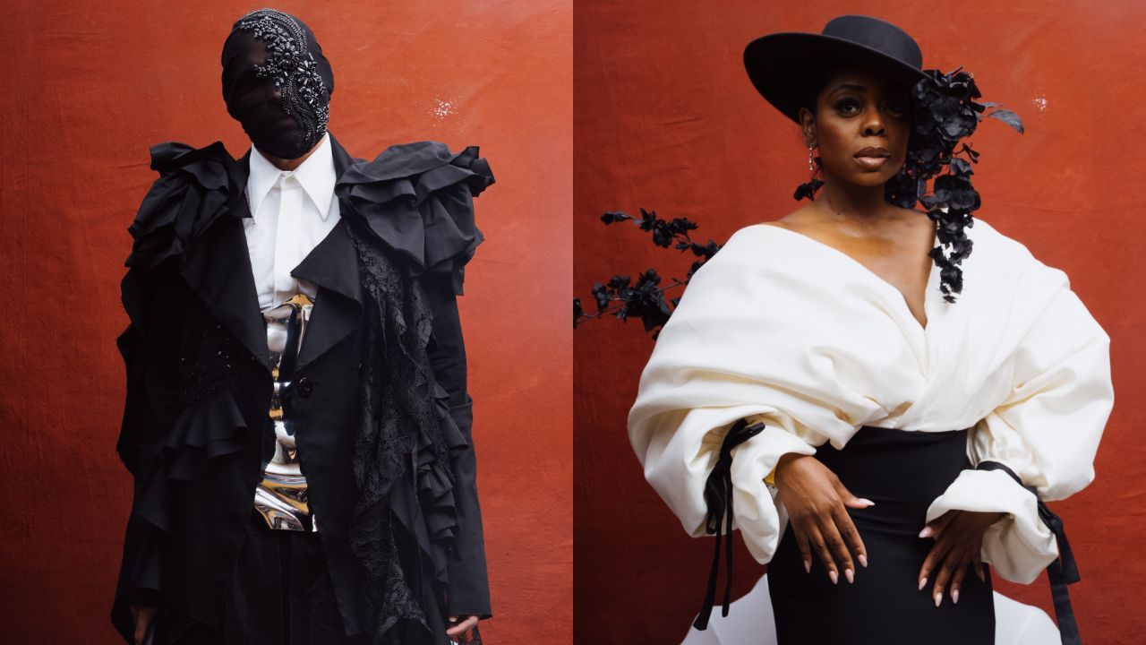 Two models pose against a red backdrop: left in a black ruffled suit with a beaded mask; right in a white oversized top, black skirt, and a wide black hat with a flower.