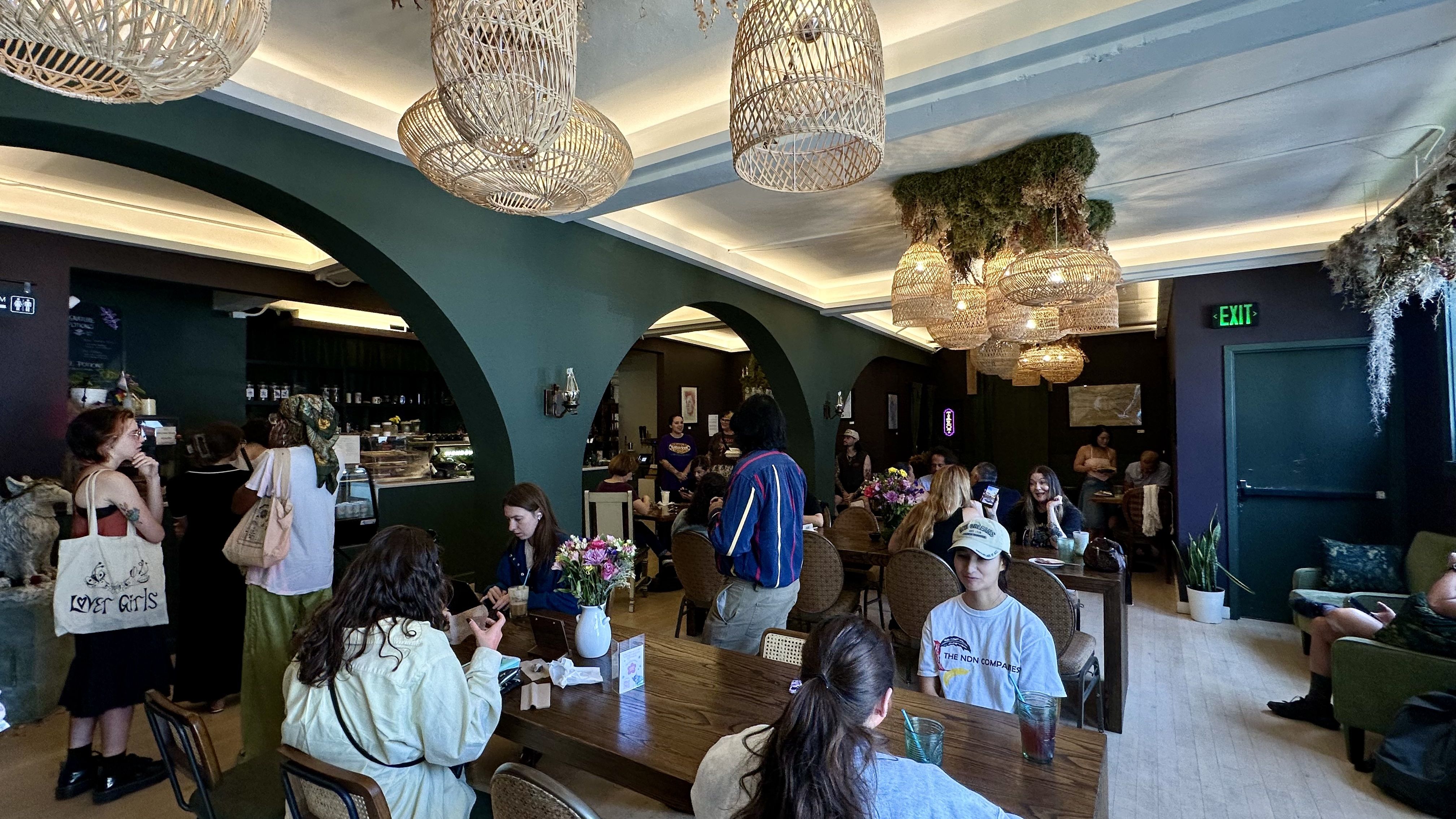 Busy cafe with green arched walls, woven wicker pendant lights, and hanging plants. People sit at wooden tables, some stand near the counter; a green EXIT sign is visible.
