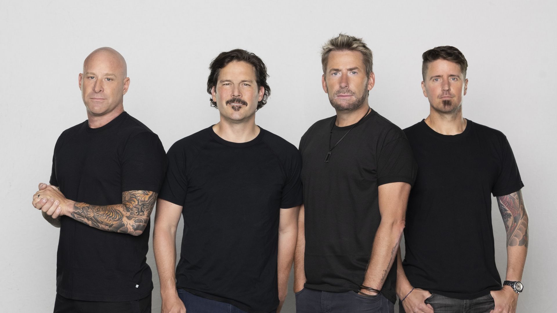 The four members of Nickelback standing against a white background, all wearing black T-shirts and casual pants, with two showing full arm tattoos and different casual footwear styles.