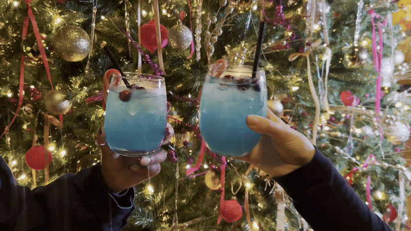 Two hands reach toward each other in a toast while they hold matching stemless wine glasses filled with a blue cocktail.