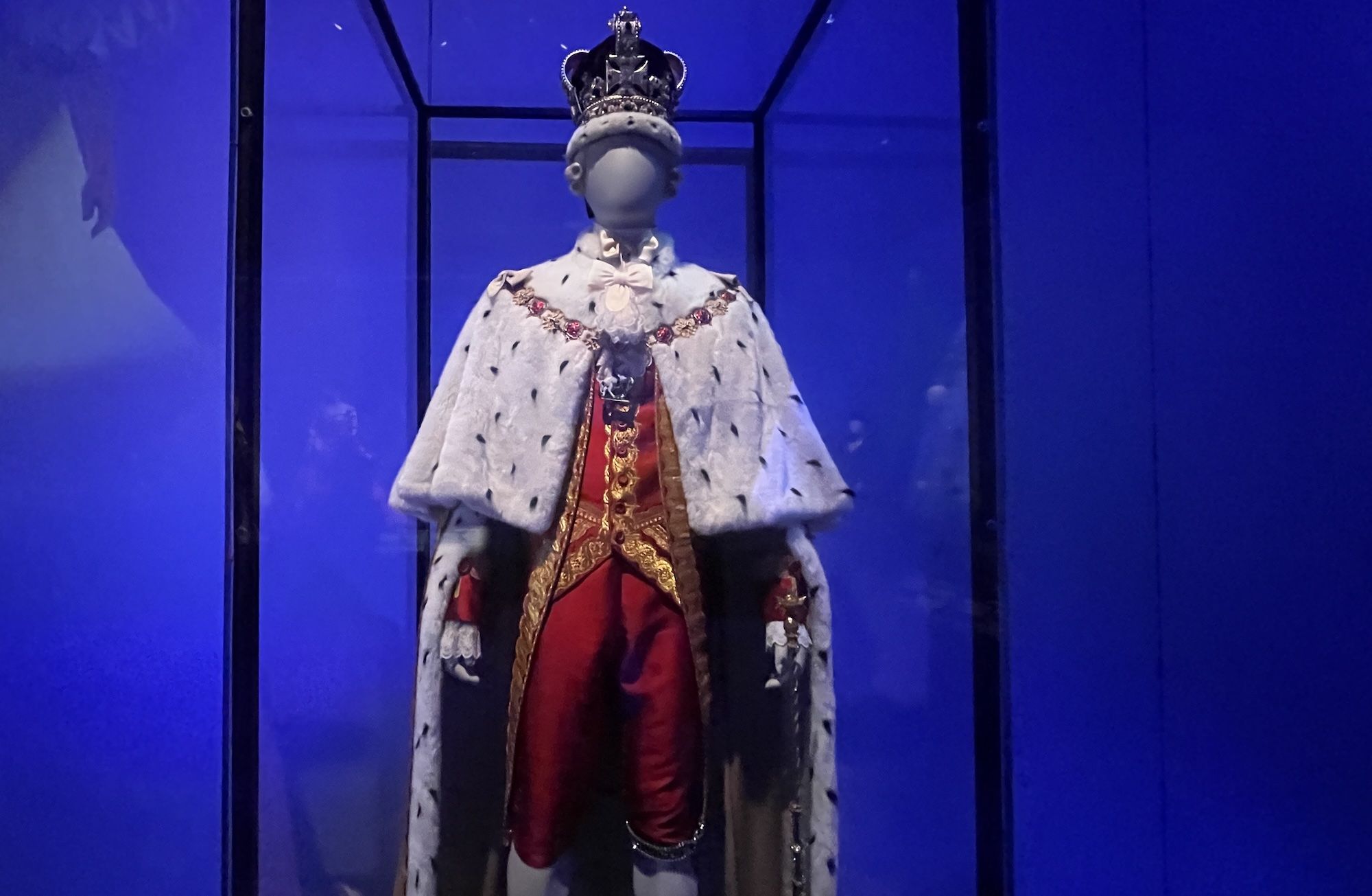 Mannequin in royal attire including a jeweled crown, white fur cape with black spots, and red and gold embellished clothing, displayed in a glass case with blue lighting.