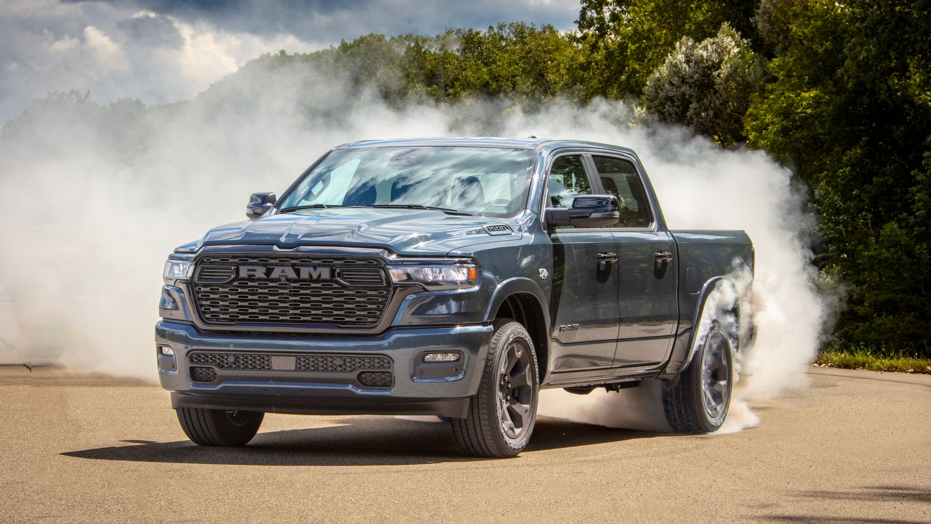 Gray Ram 1500 Hemi pickup truck kicking up dust on a dirt road, with smoke billowing around the rear tires, and green trees in the background under a cloudy sky.