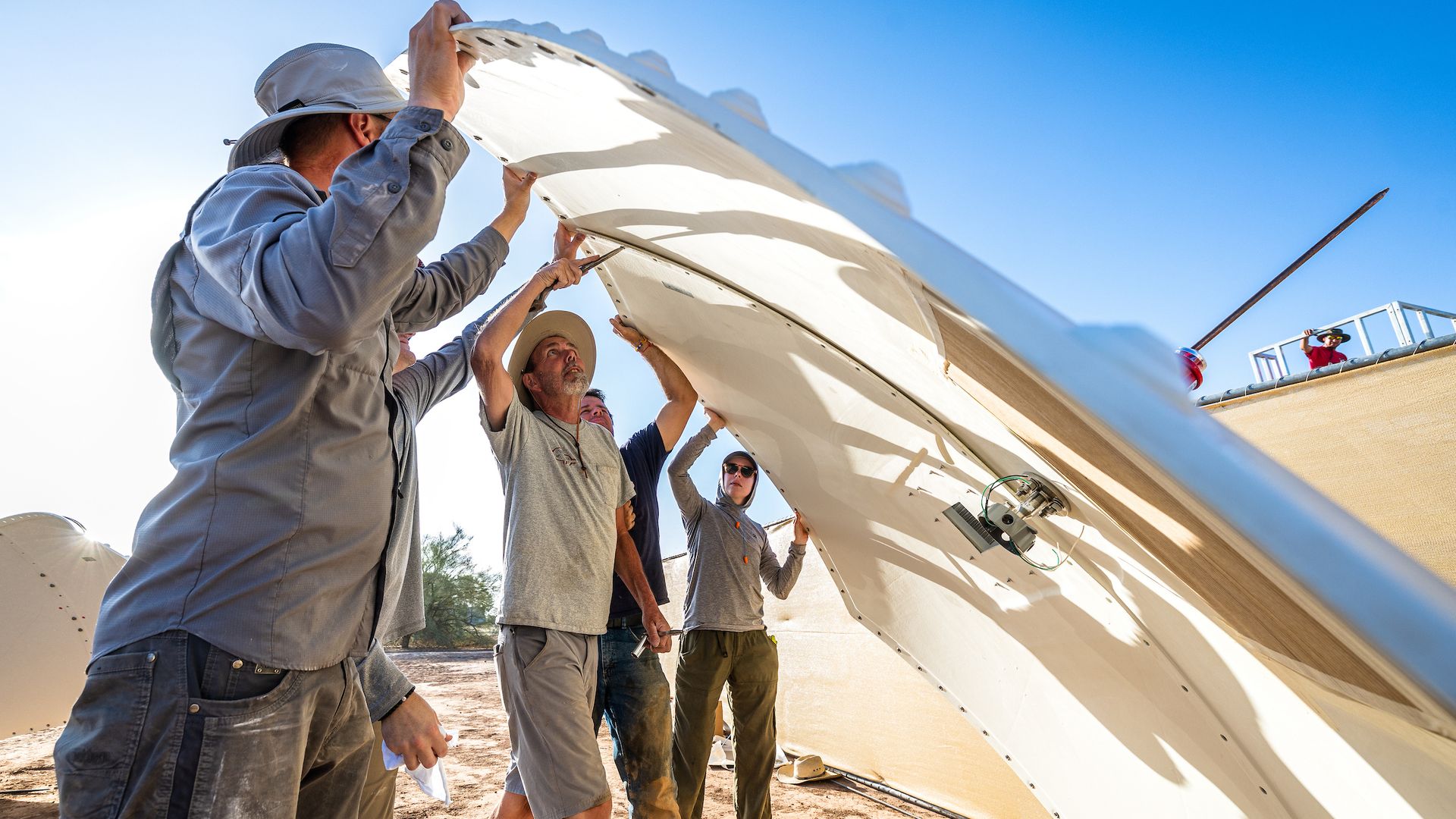 Group of workers in gray shirts and hats lift and trim a large white aircraft wing panel outdoors, under a bright blue sky with dusty ground.