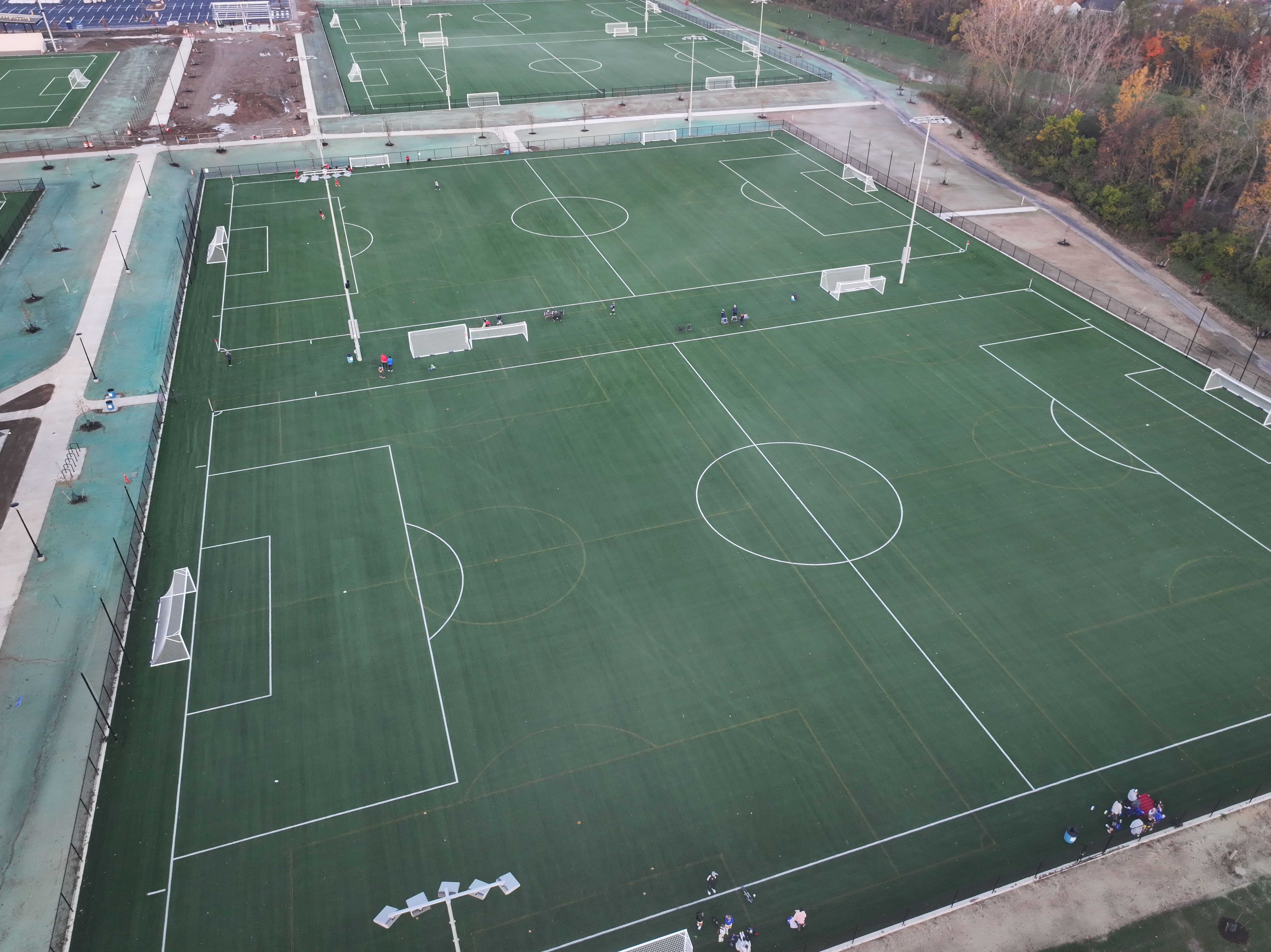 Aerial view of multiple green artificial turf soccer fields with white goalposts and markings, a few people scattered along the sidelines, and trees in the background.