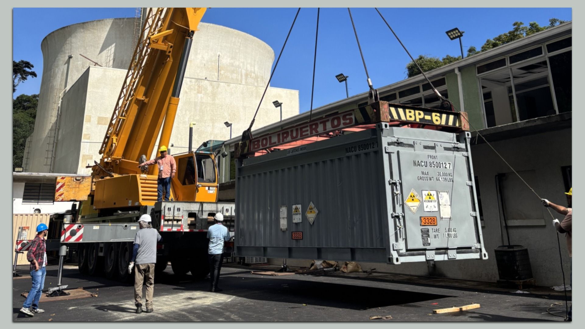 Yellow crane lifts a gray shipping container onto a trailer. Workers in hard hats watch, with a circular concrete tank and a building in the background.