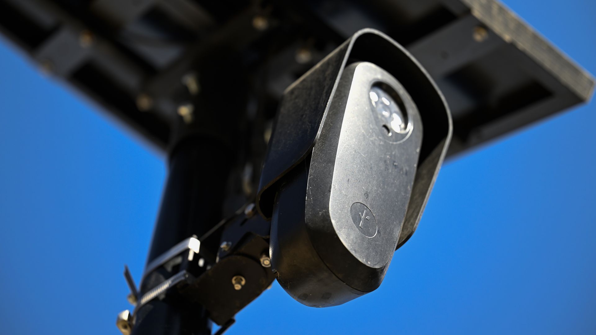Close-up of a dark metal hinge/arm with curved plates and visible bolts, part of heavy machinery, set against a bright blue sky.